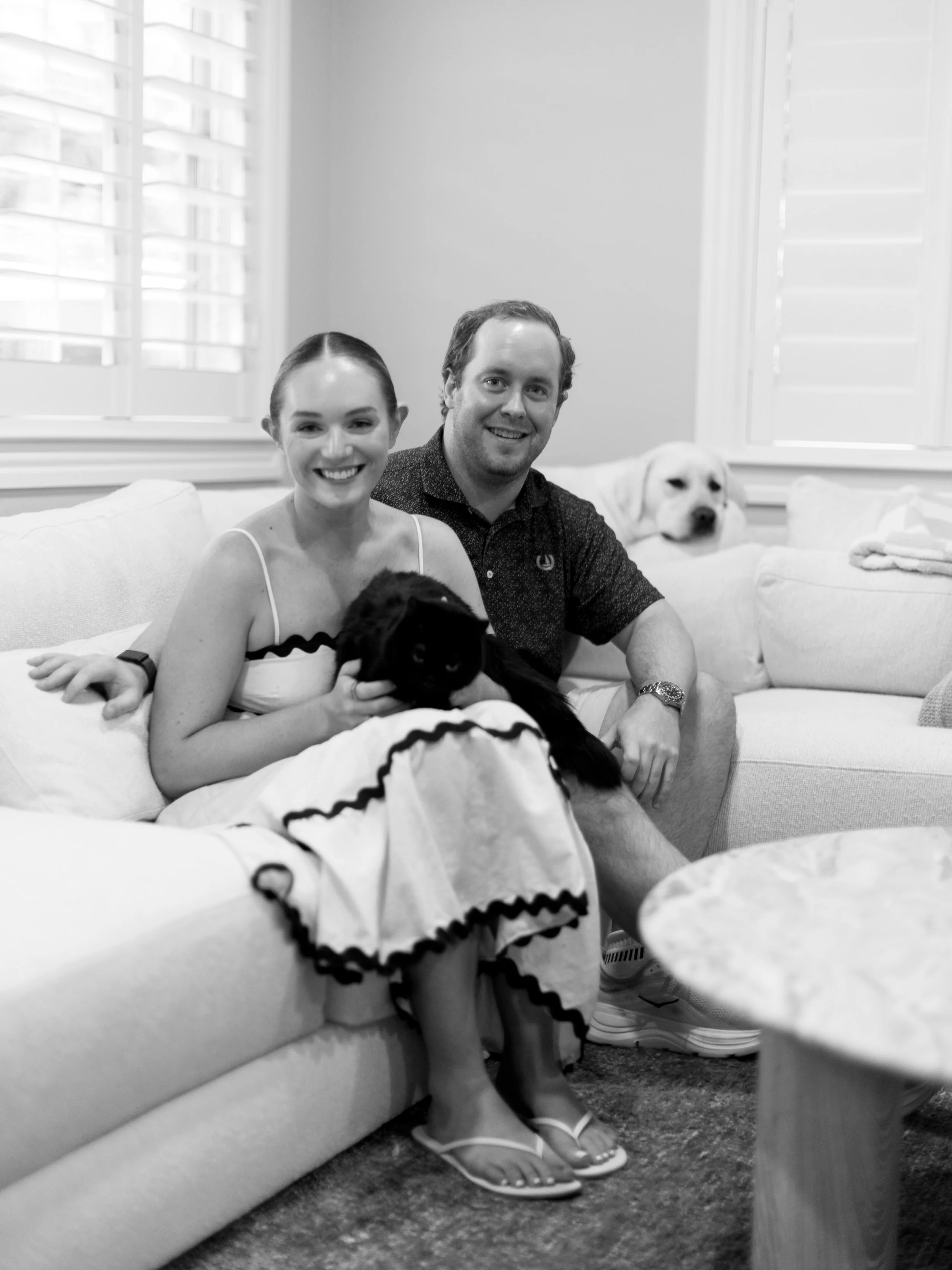A smiling woman and man sitting on a sofa with two dogs, one black and one white, in a living room with bright windows and curtains.