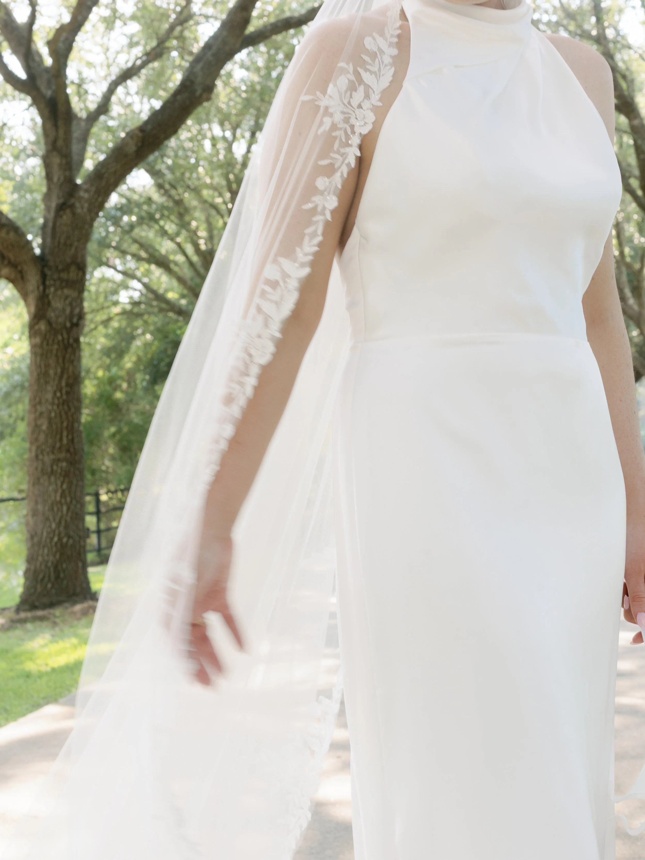 Close-up of a bride wearing a white wedding dress with embroidered lace details on the shoulder and a sheer veil, standing outdoors among trees.