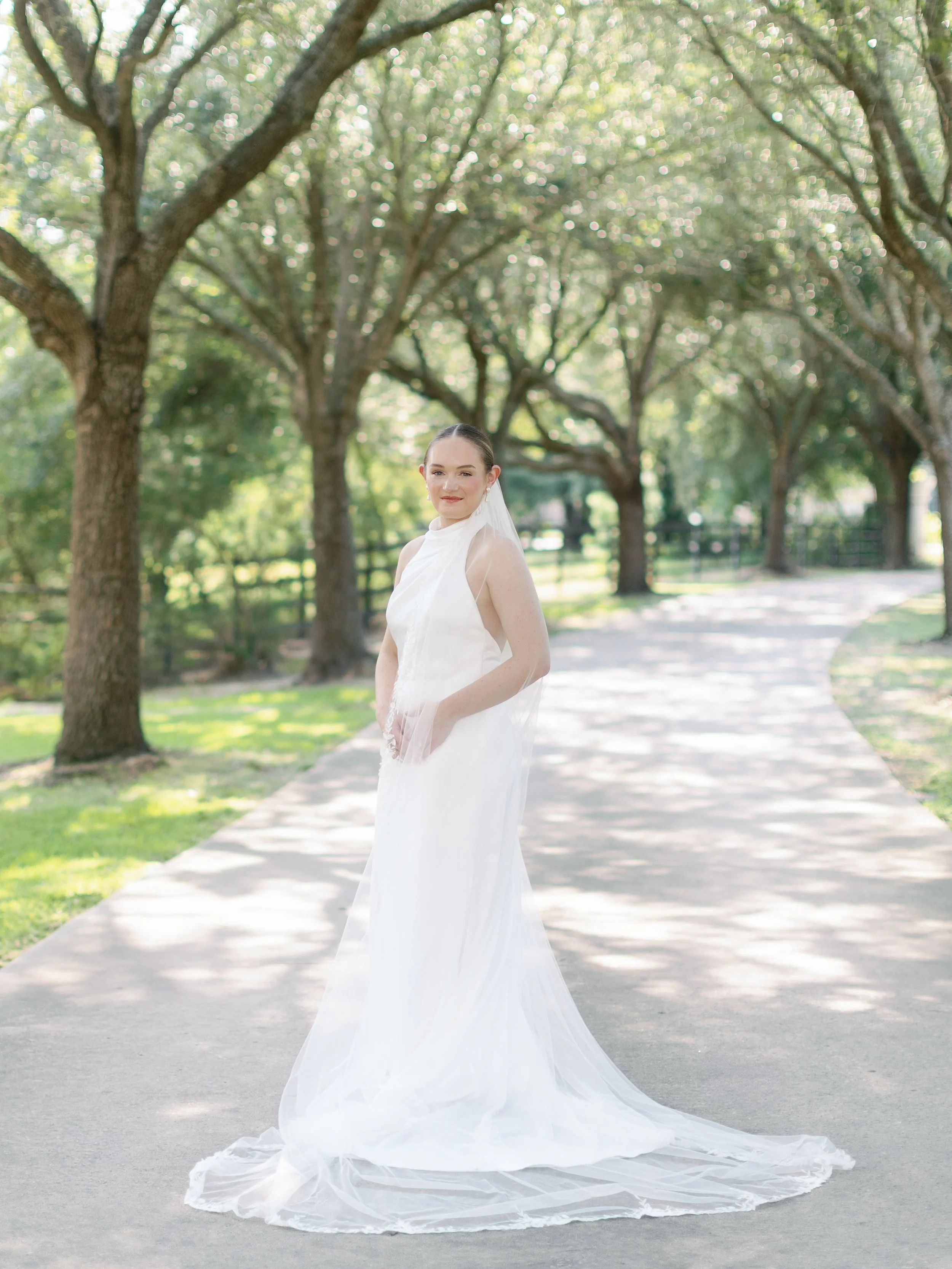 A woman in a white wedding gown with a veil stands on a paved path surrounded by trees with green leaves, in a park or garden setting, during daytime.