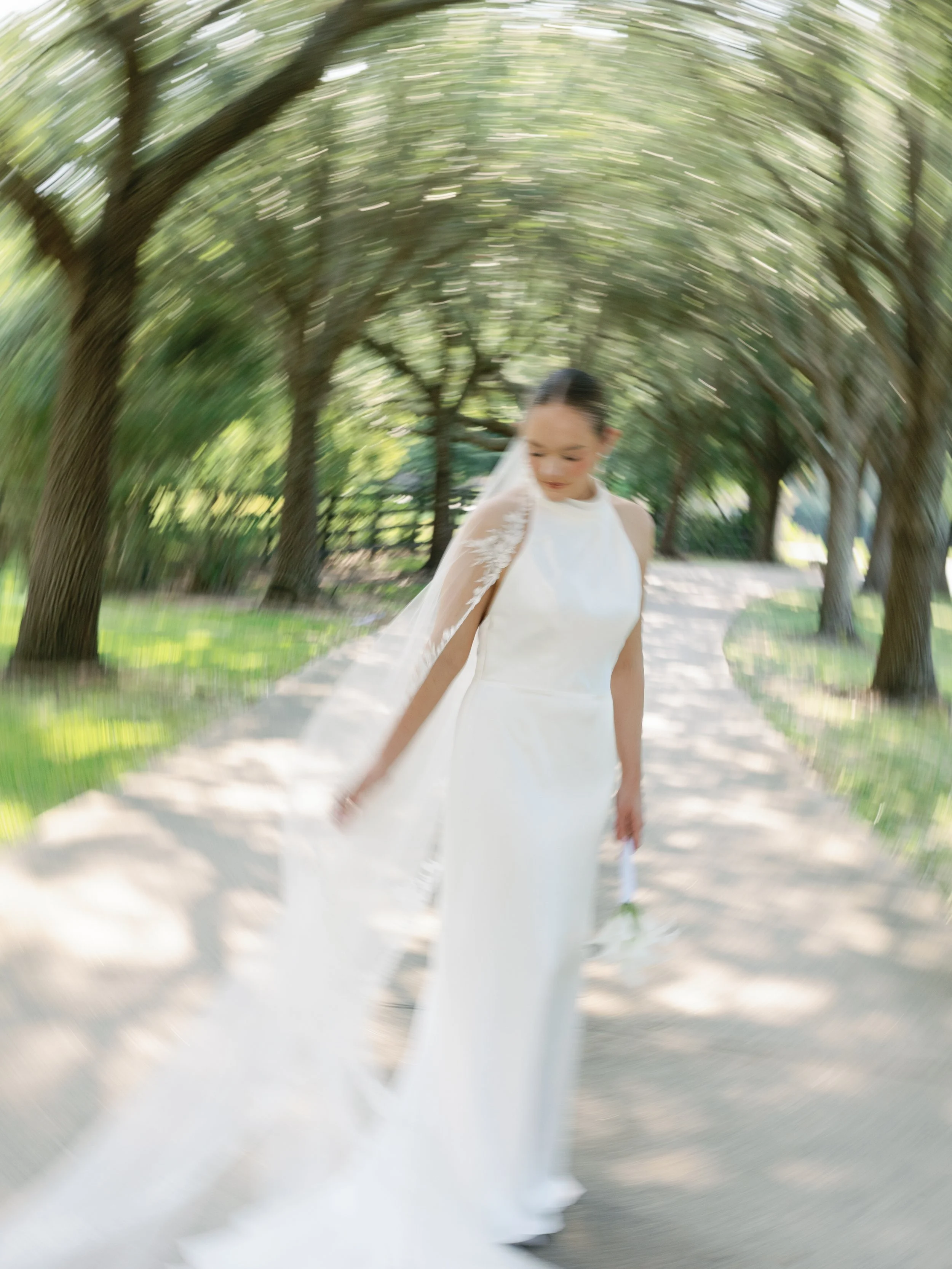 A bride in a white wedding dress with a lace veil walking on a tree-lined path.