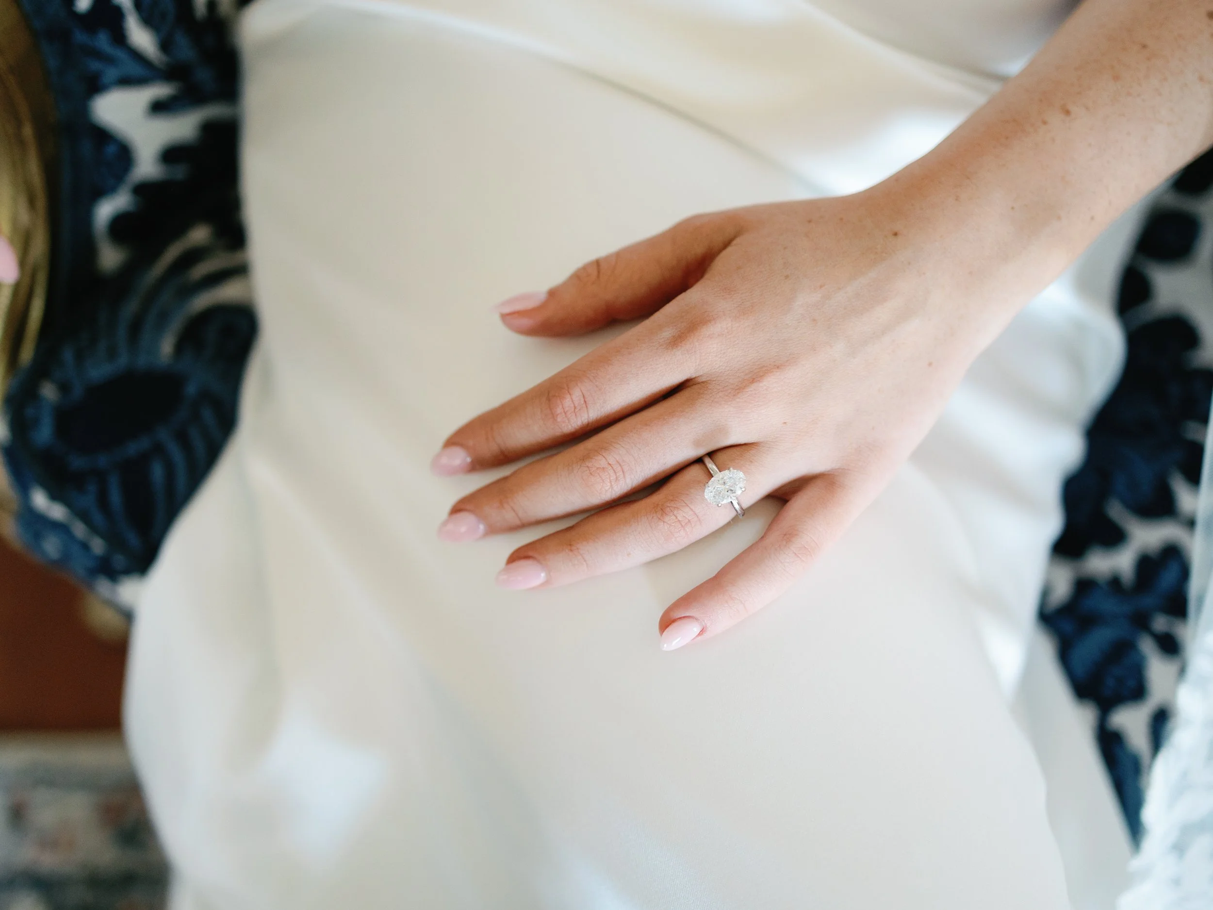 Close-up of a woman's hand with a large diamond ring on her ring finger, resting on a white fabric surface.
