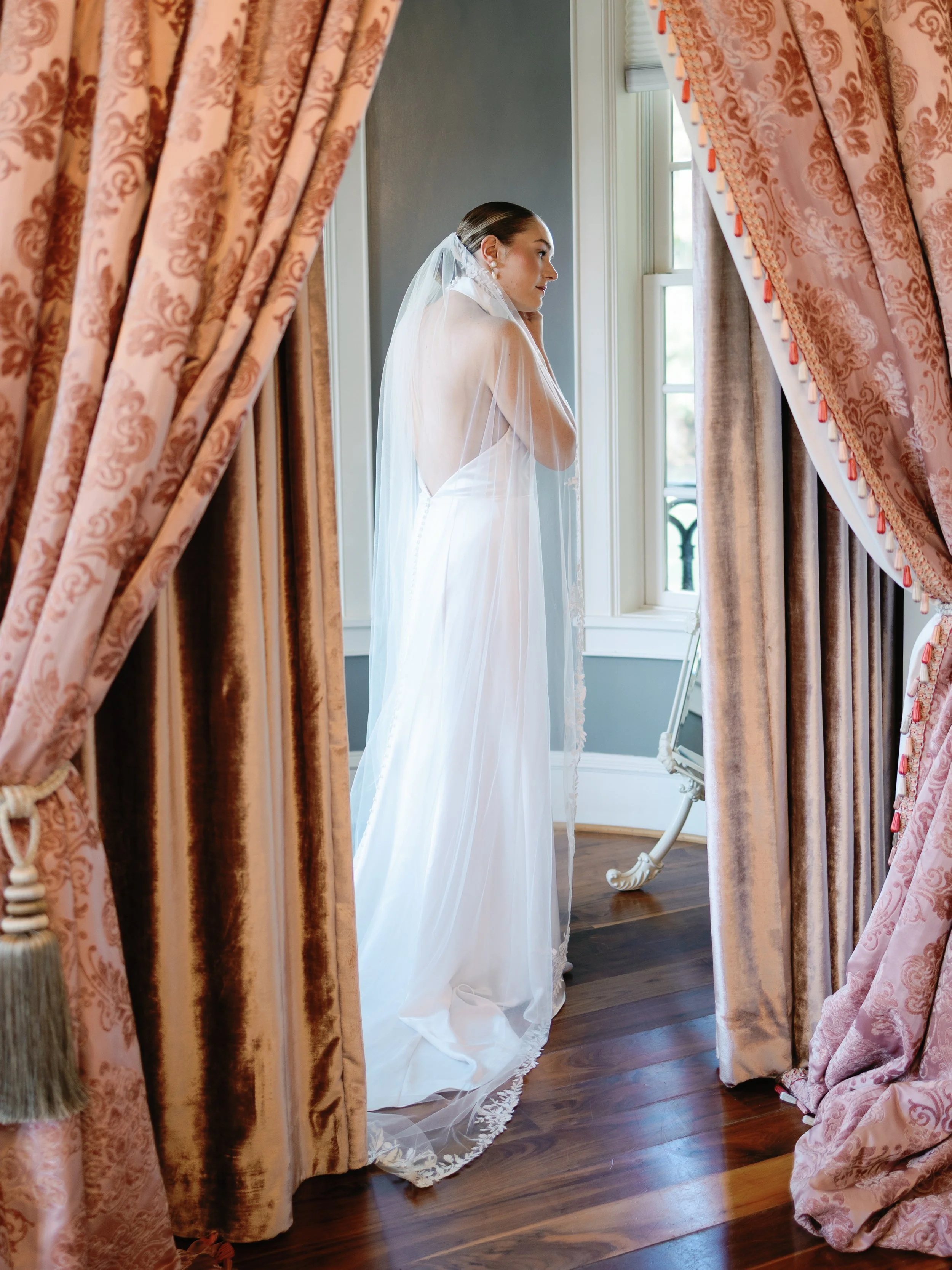 A bride in a white wedding gown with a long veil, standing by a window, appearing to be in a quiet, reflective moment.