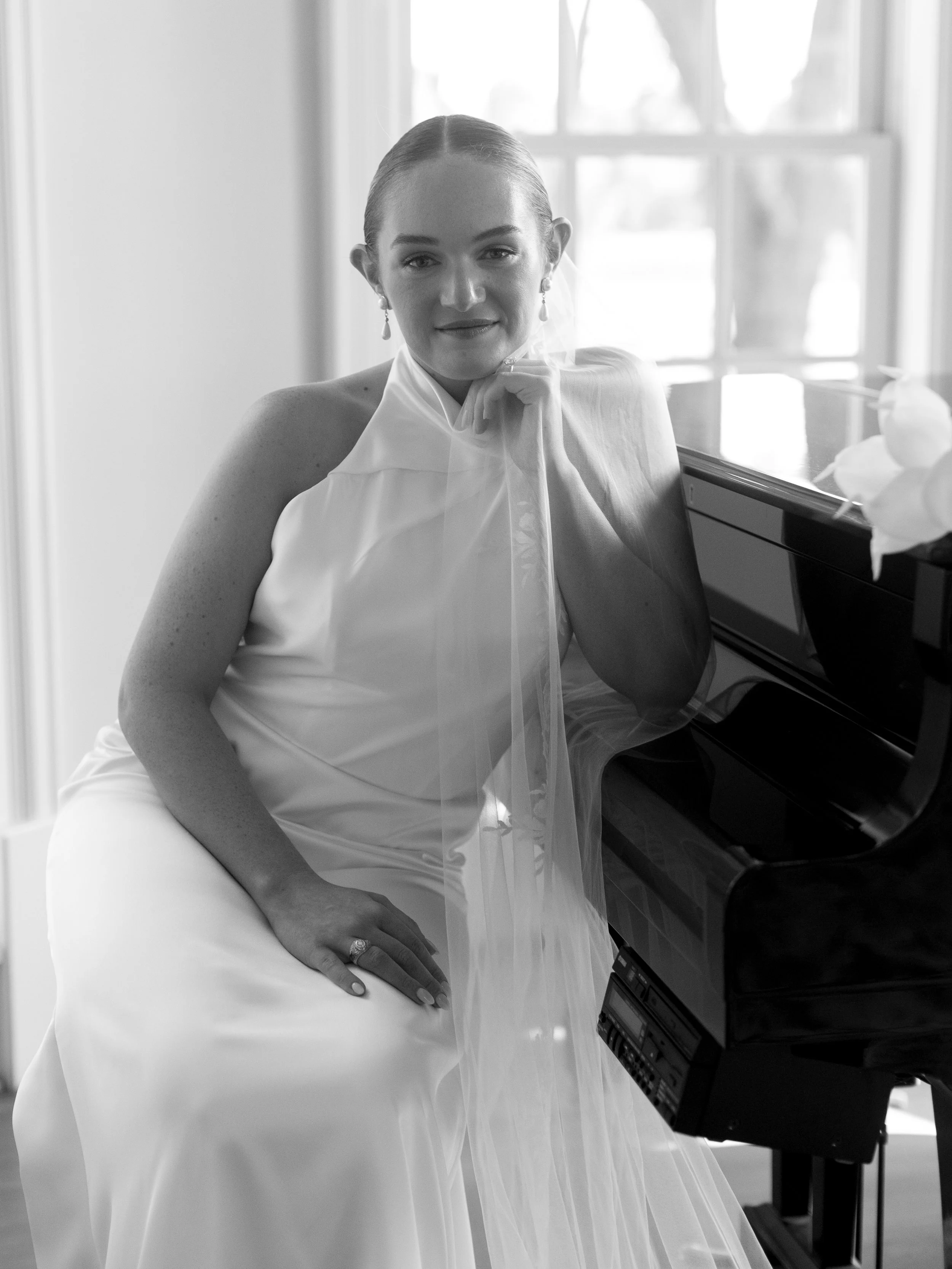 A woman in a bridal gown sitting at a piano, with her chin resting on her hand, smiling softly.