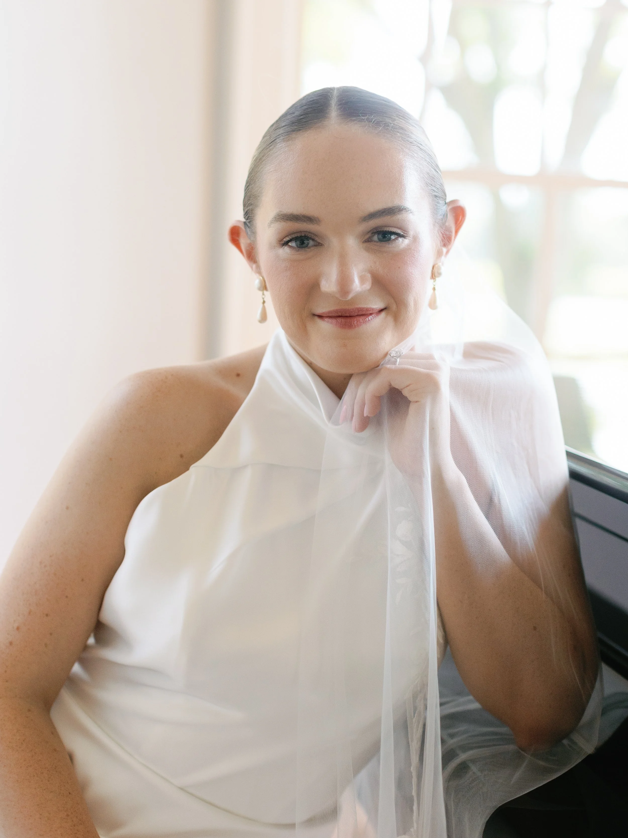 Portrait of a woman with slicked-back hair, wearing elegant earrings and a white sleeveless dress, smiling softly, and resting her chin on her hand with sheer fabric draped over her shoulder, sitting near a window with a blurred outdoor background.