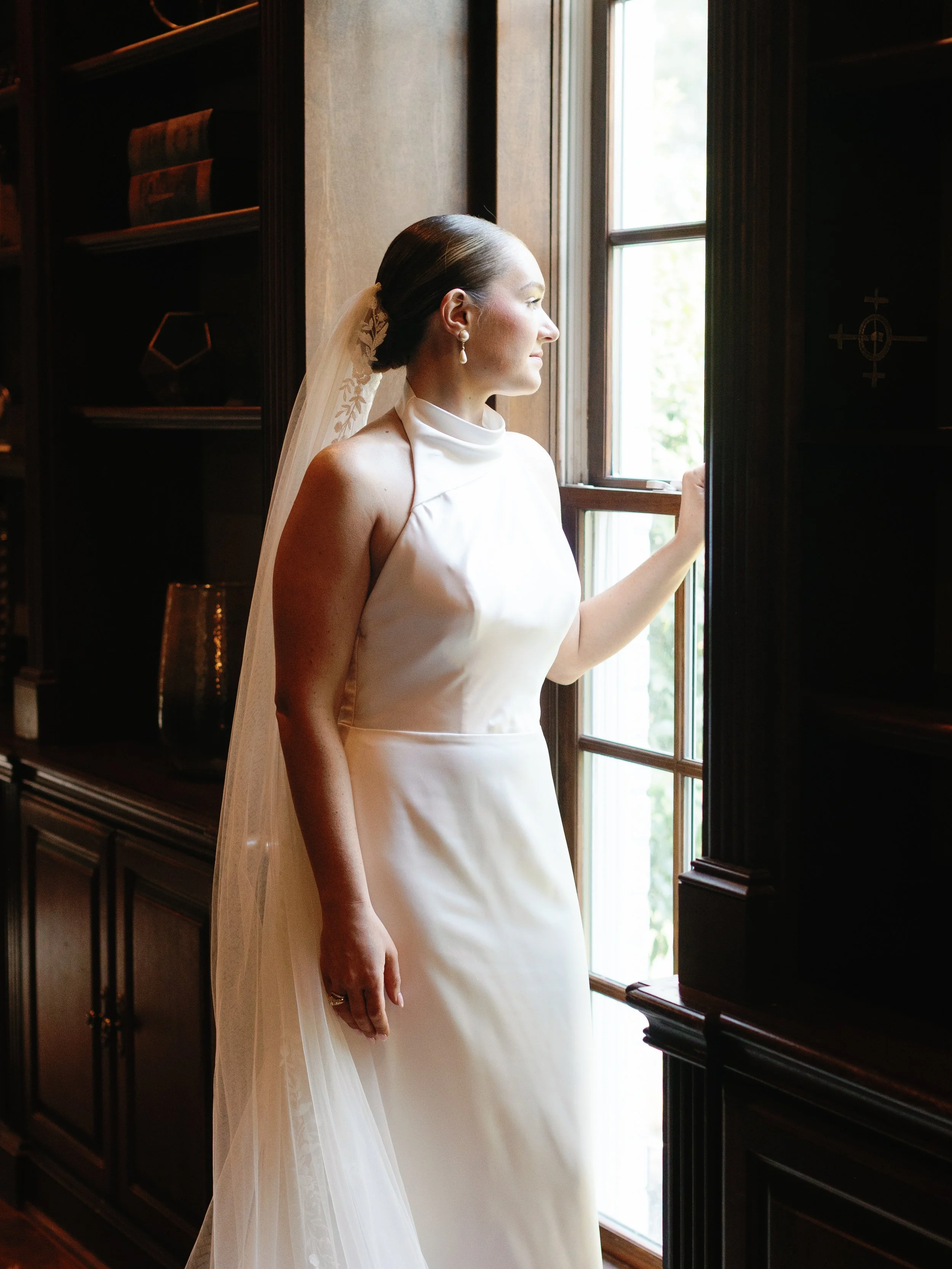 Bridal woman in white dress looking out of a window in a wood-paneled room.