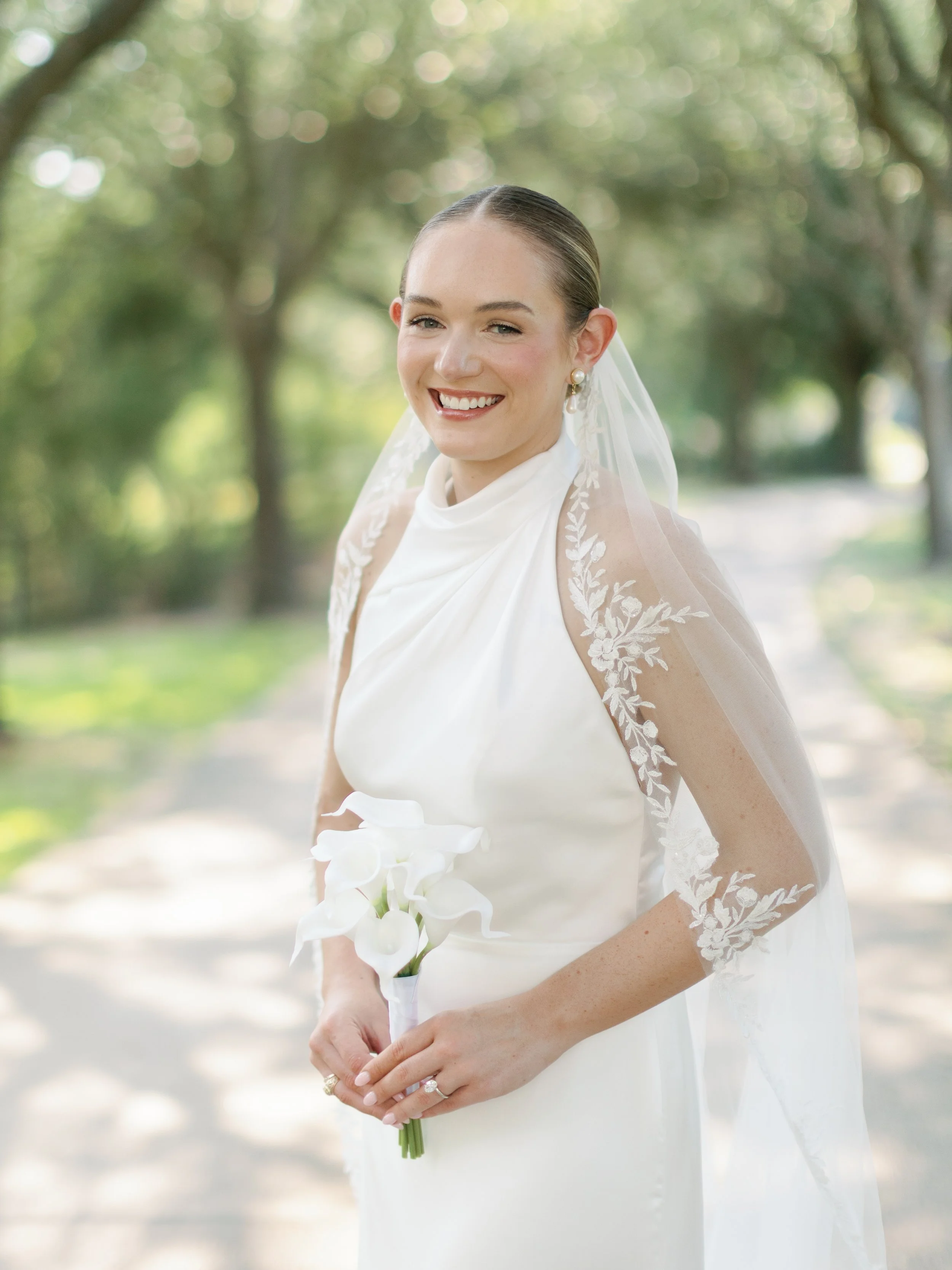 A smiling bride in a white wedding dress holding a bouquet of white calla lilies on a sunlit outdoor path with trees in the background.
