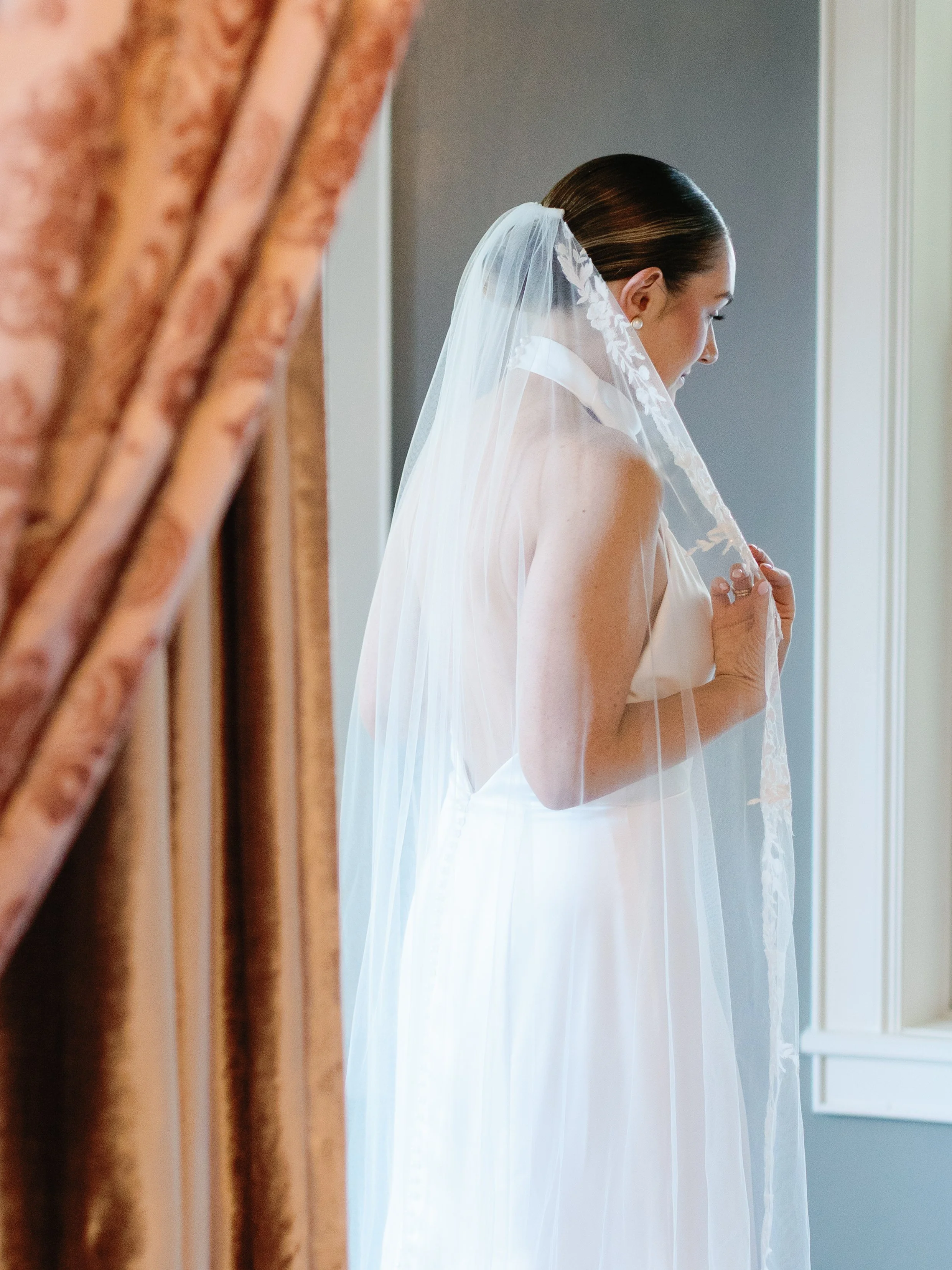 A bride in a white wedding gown with a lace veil is standing by a window, adjusting her veil.