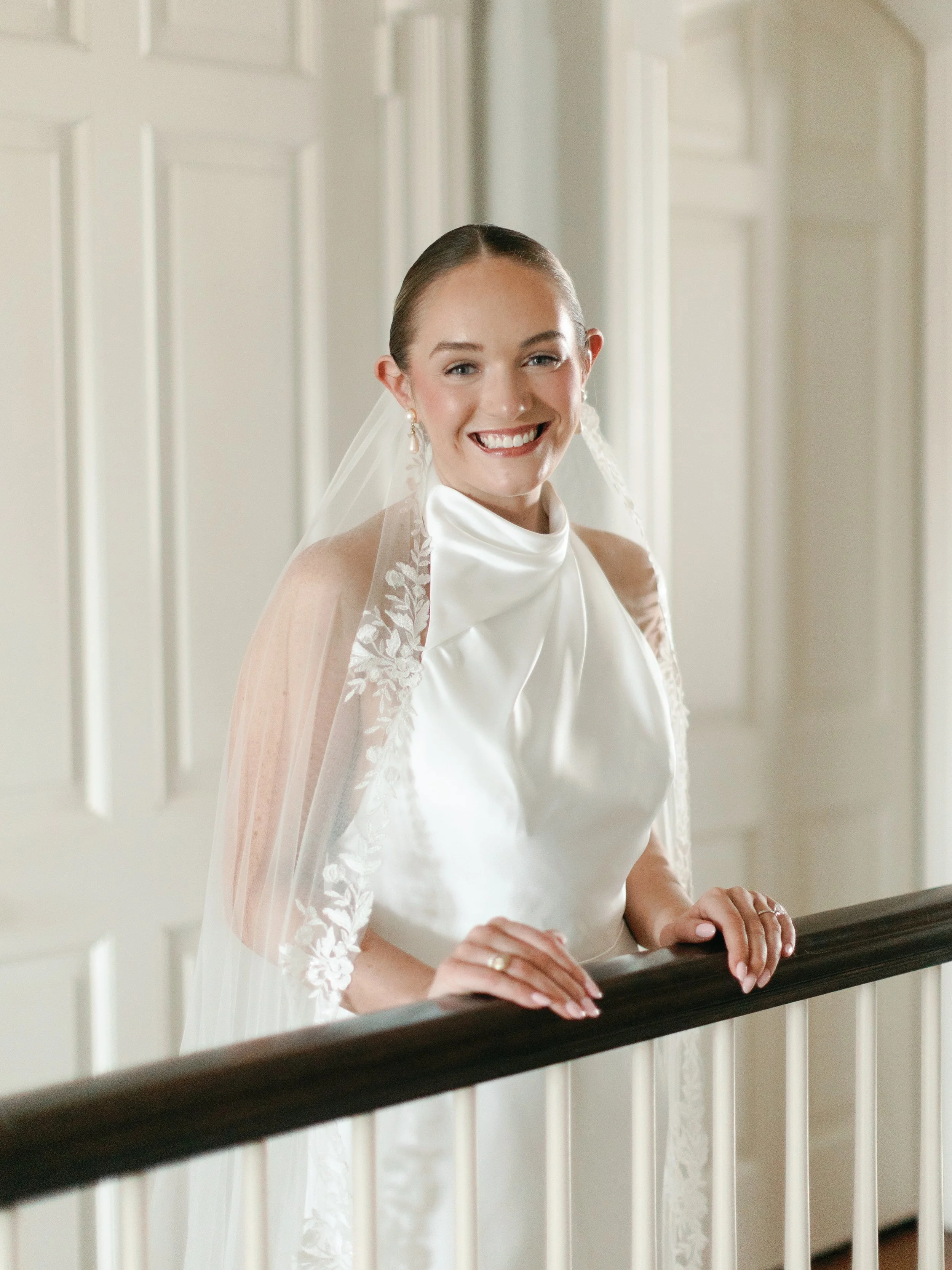 A smiling bride in a white wedding gown with a veil, standing indoors on a staircase with a dark wooden handrail.