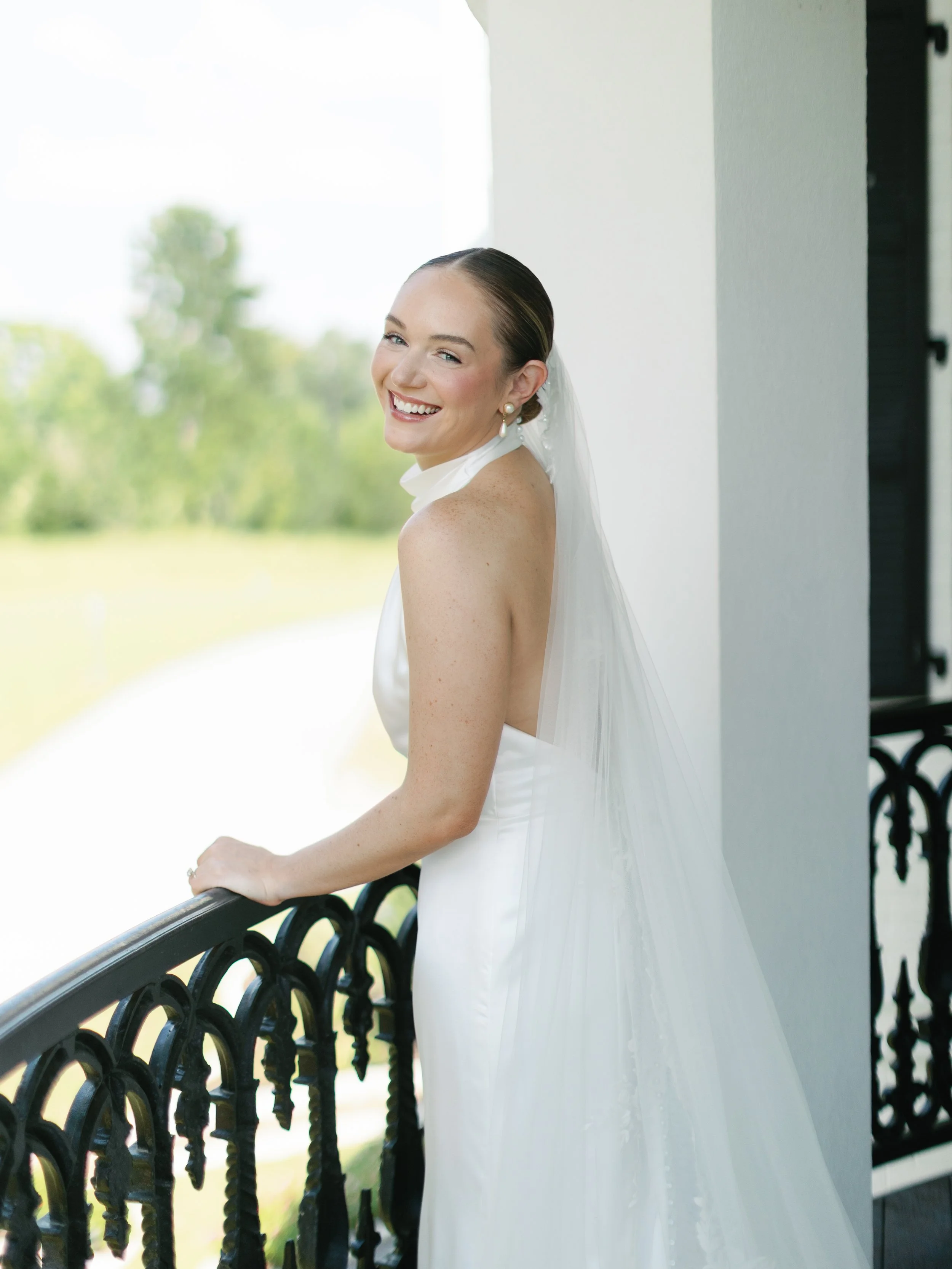 A bride standing on a balcony, smiling and looking at the camera, wearing a white wedding dress with a veil, outdoors with green trees in the background.