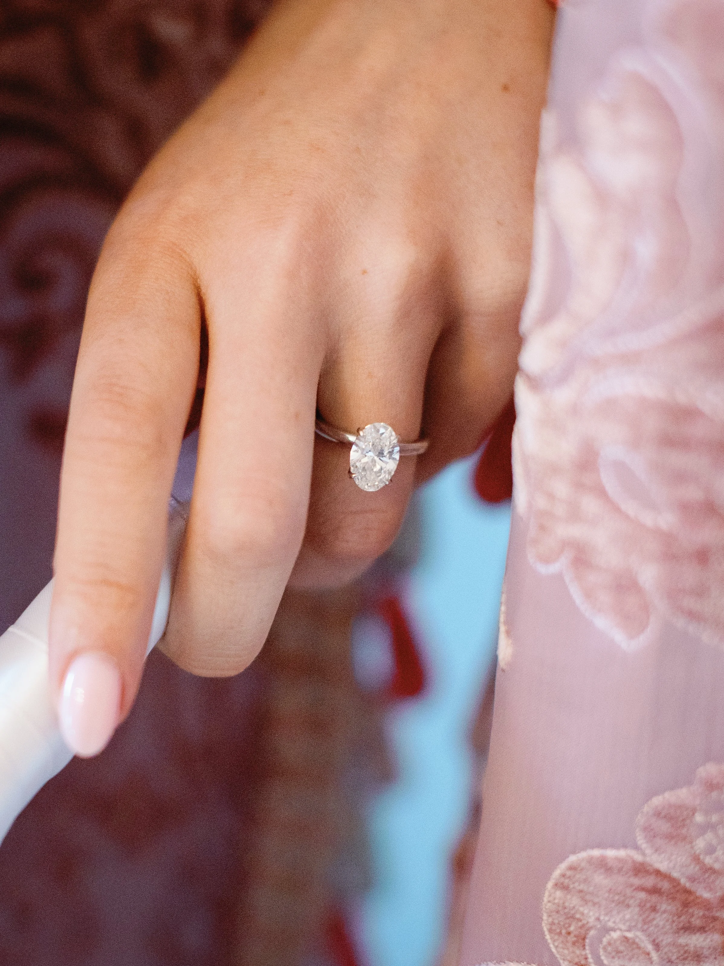 Close-up of a hand wearing a silver ring with a large oval diamond, resting on pink fabric with floral embroidery.