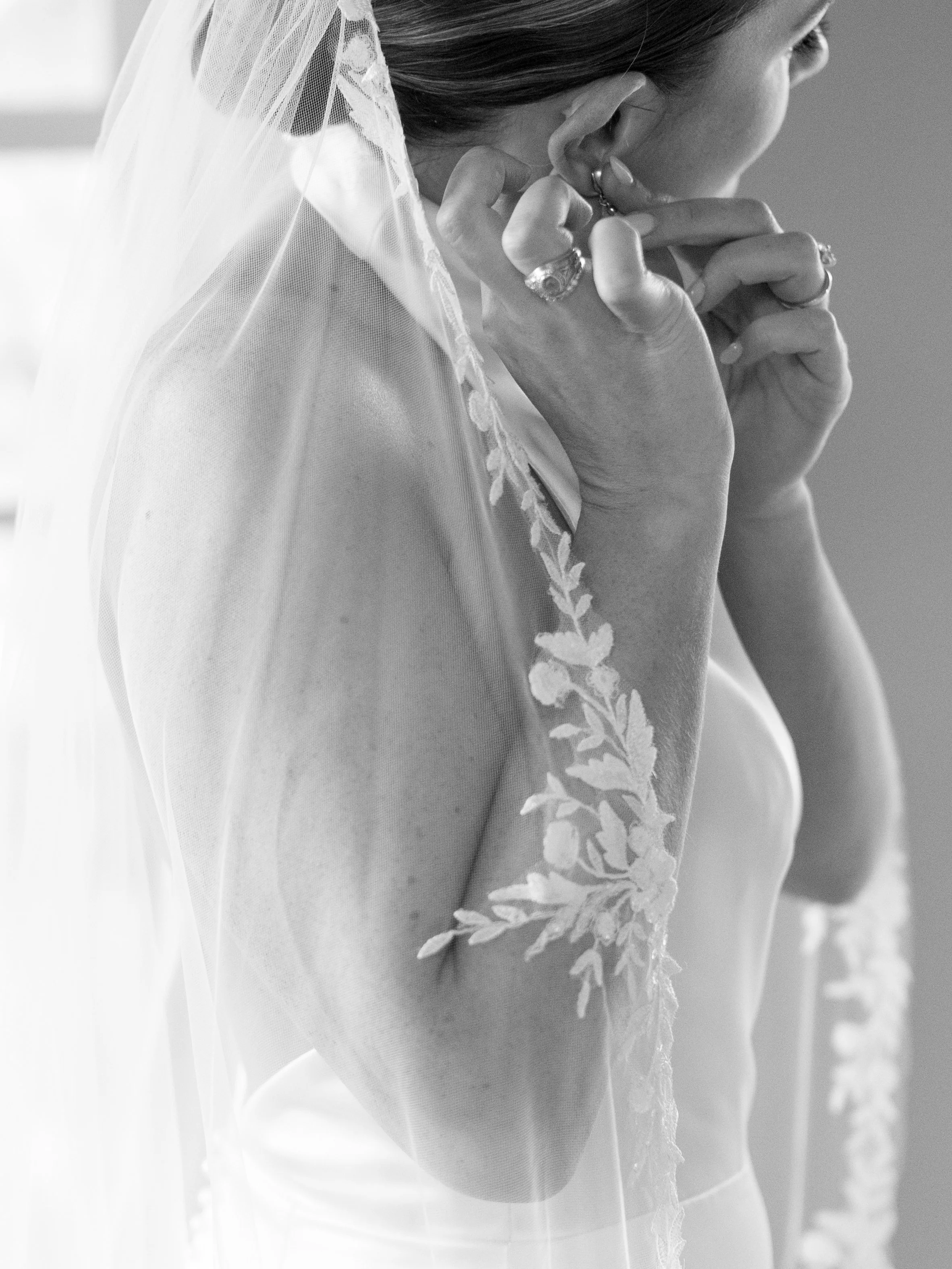 A woman in a wedding dress adjusting her earring, with a veil and lace detail on her dress.