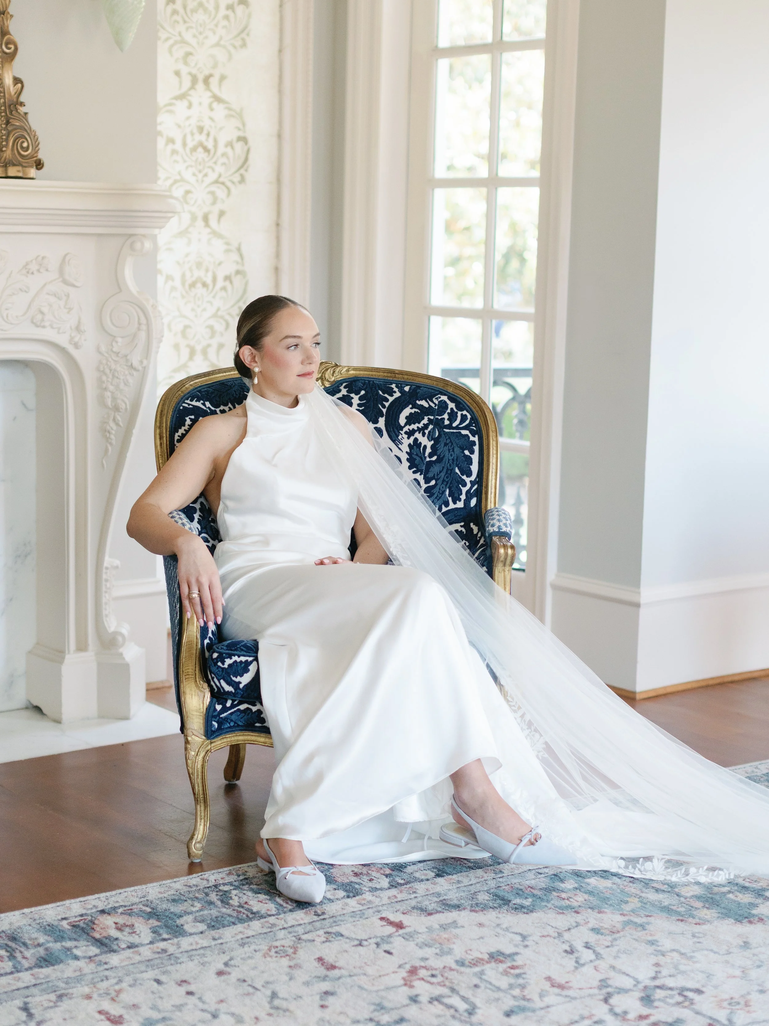 A woman in a white wedding dress with a veil, sitting on a vintage blue and gold armchair in a well-lit room with large windows and ornate decor.