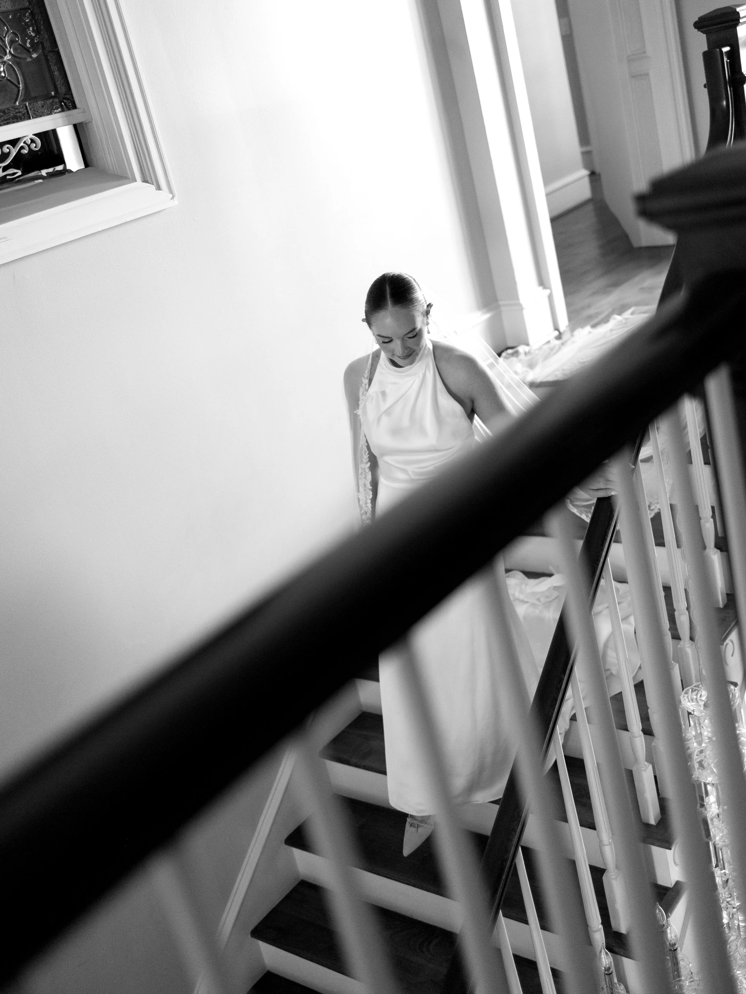 A woman in a white wedding dress standing on a tree-lined pathway in a park, smiling at the camera.