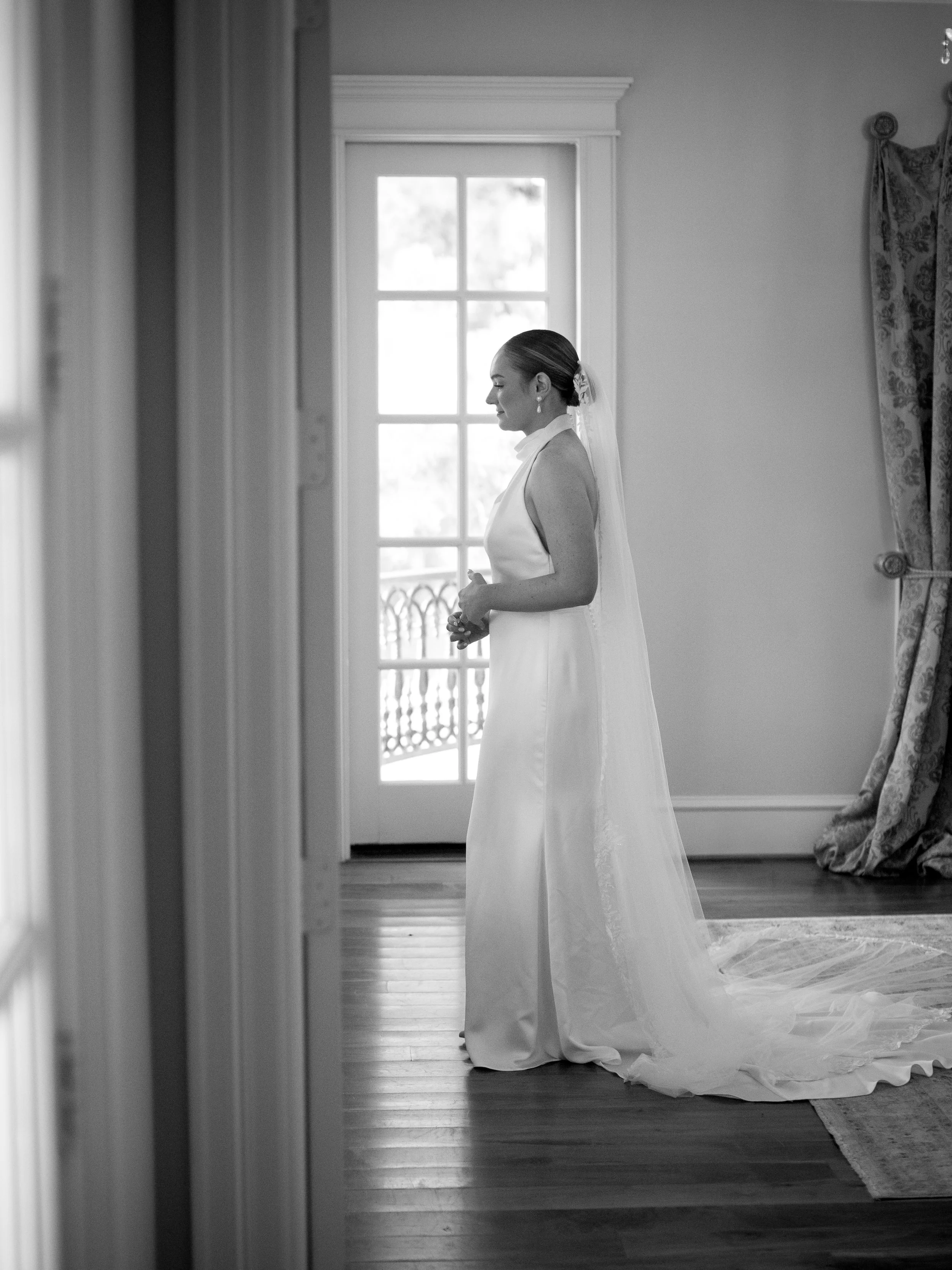Black and white photo of a bride in a wedding dress standing in a room near a window, with her hands clasped and a veil on her hair.