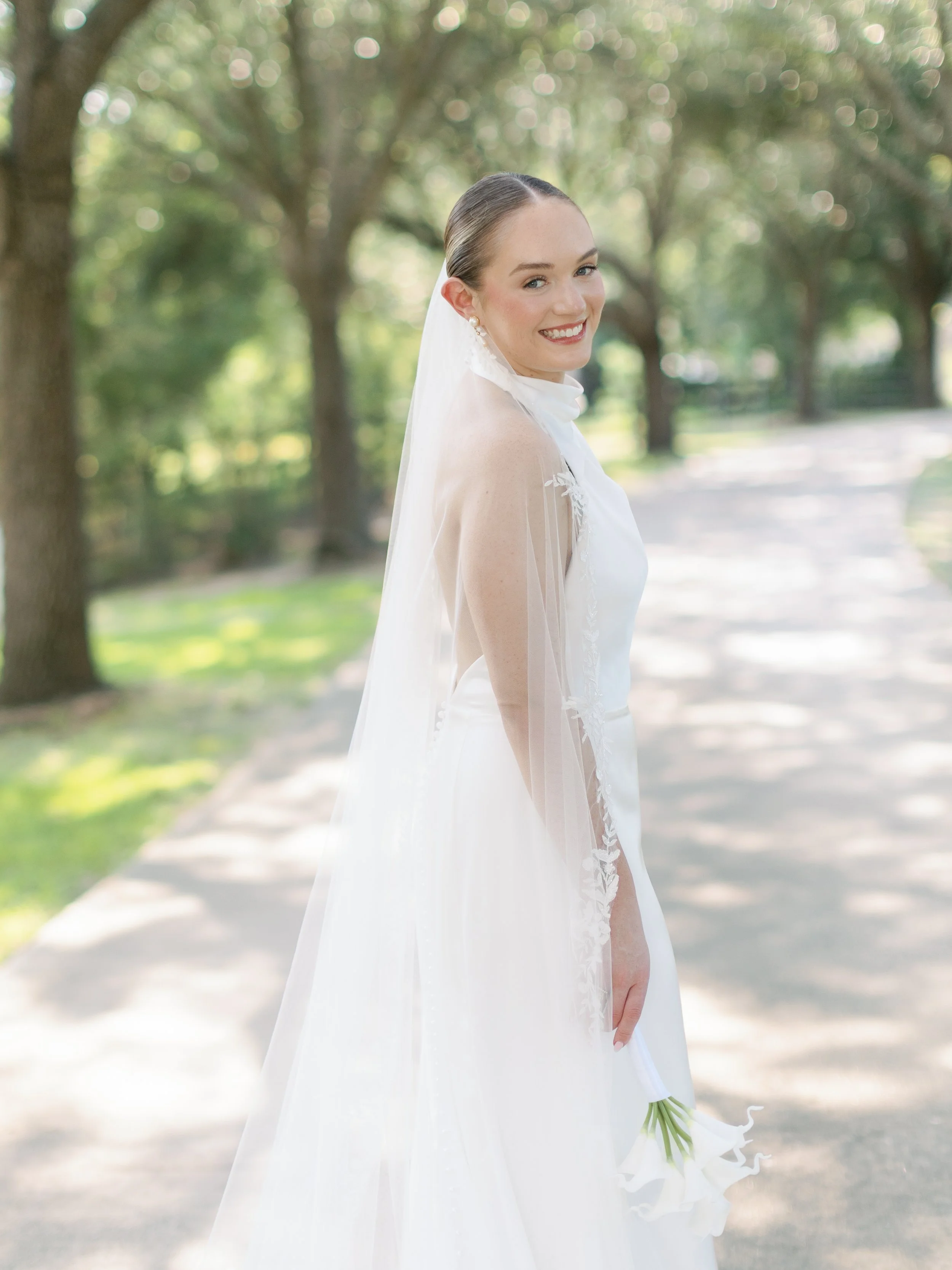 A bride in a white wedding dress with a veil, holding white flowers, smiling outdoors on a sunny day.
