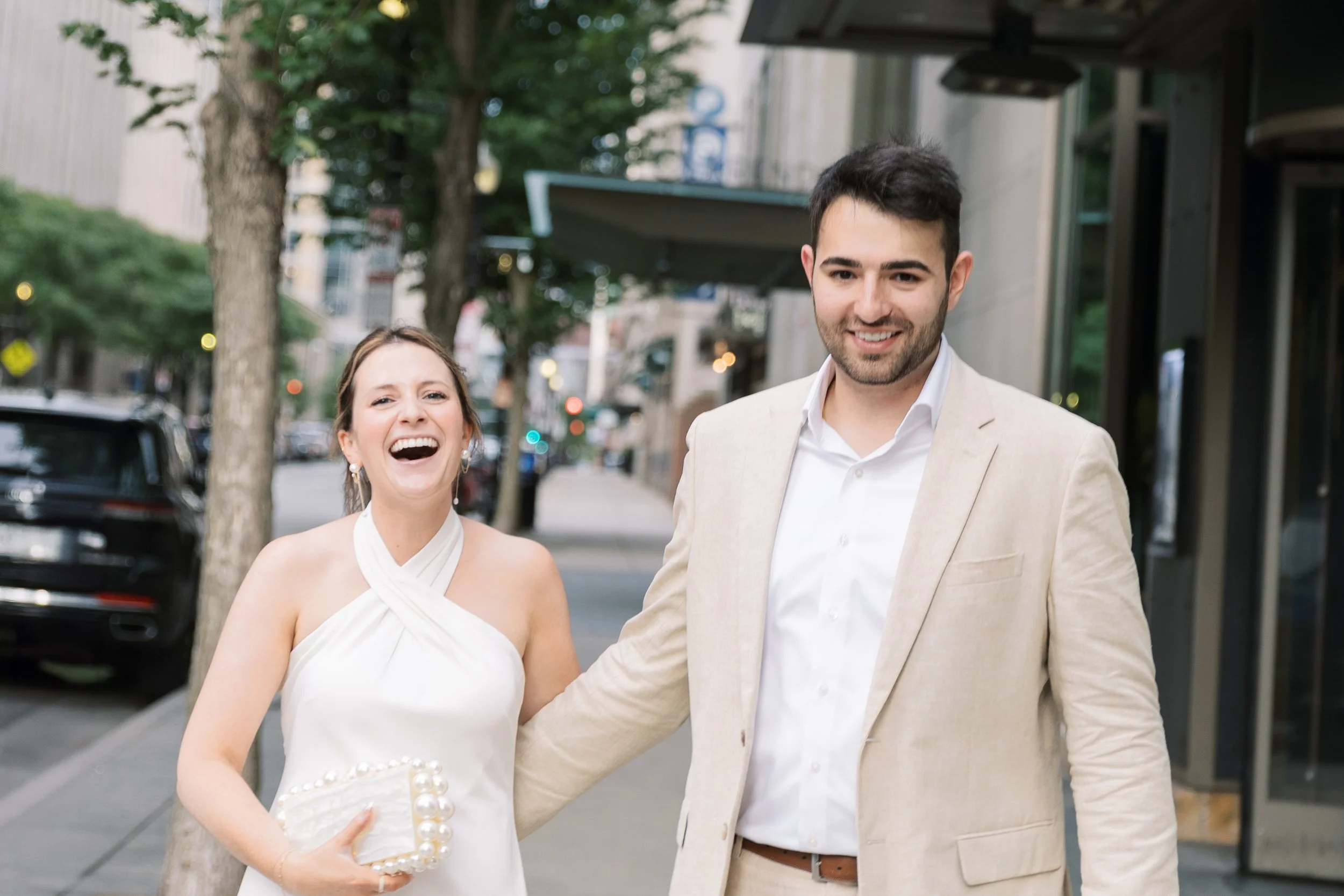A smiling young woman in a white dress and young man in a beige suit walking outdoors on a city sidewalk, with trees and parked cars in the background.