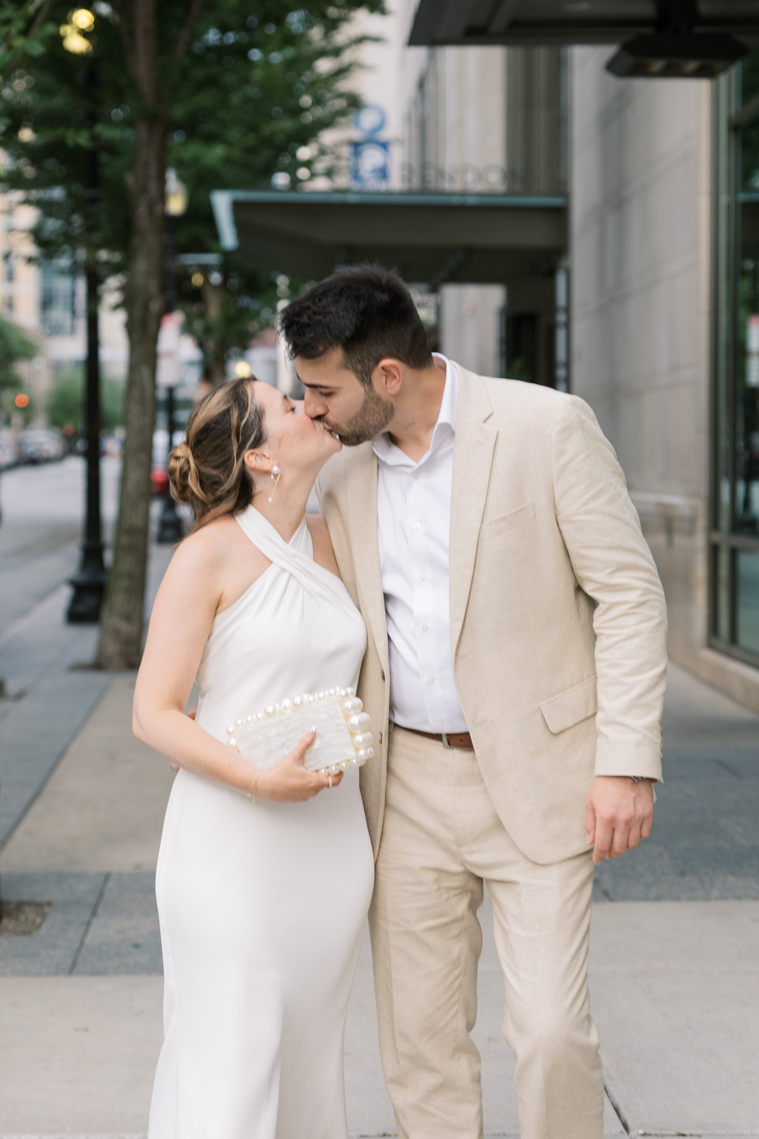 A newlywed couple sharing a kiss on a city street during the daytime. The bride is wearing a white wedding dress and holding a clutch, while the groom is dressed in a beige suit jacket and matching trousers.