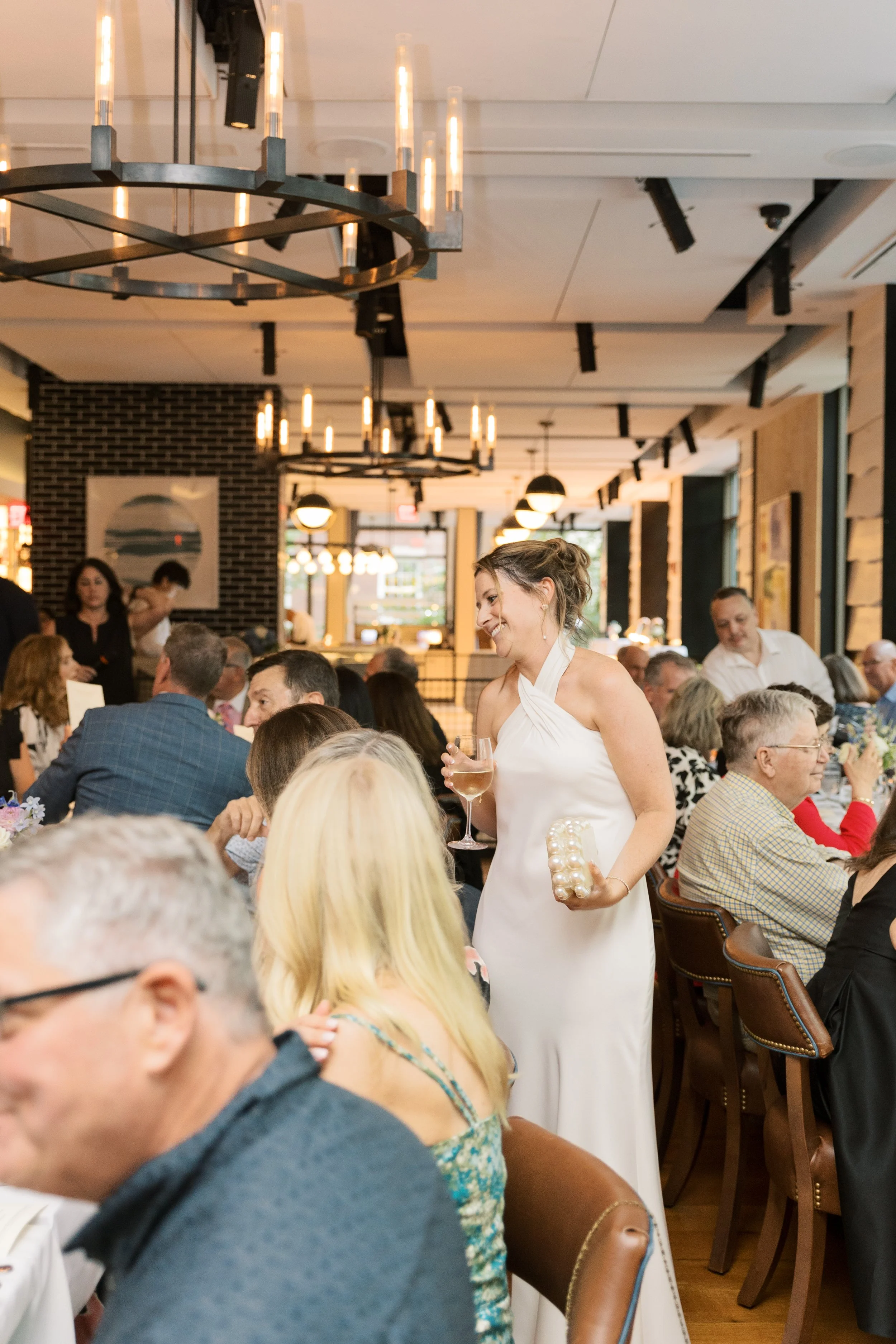 A woman in a white dress holding a glass of wine and a small gift box, standing and smiling at a social gathering in a restaurant or banquet hall.