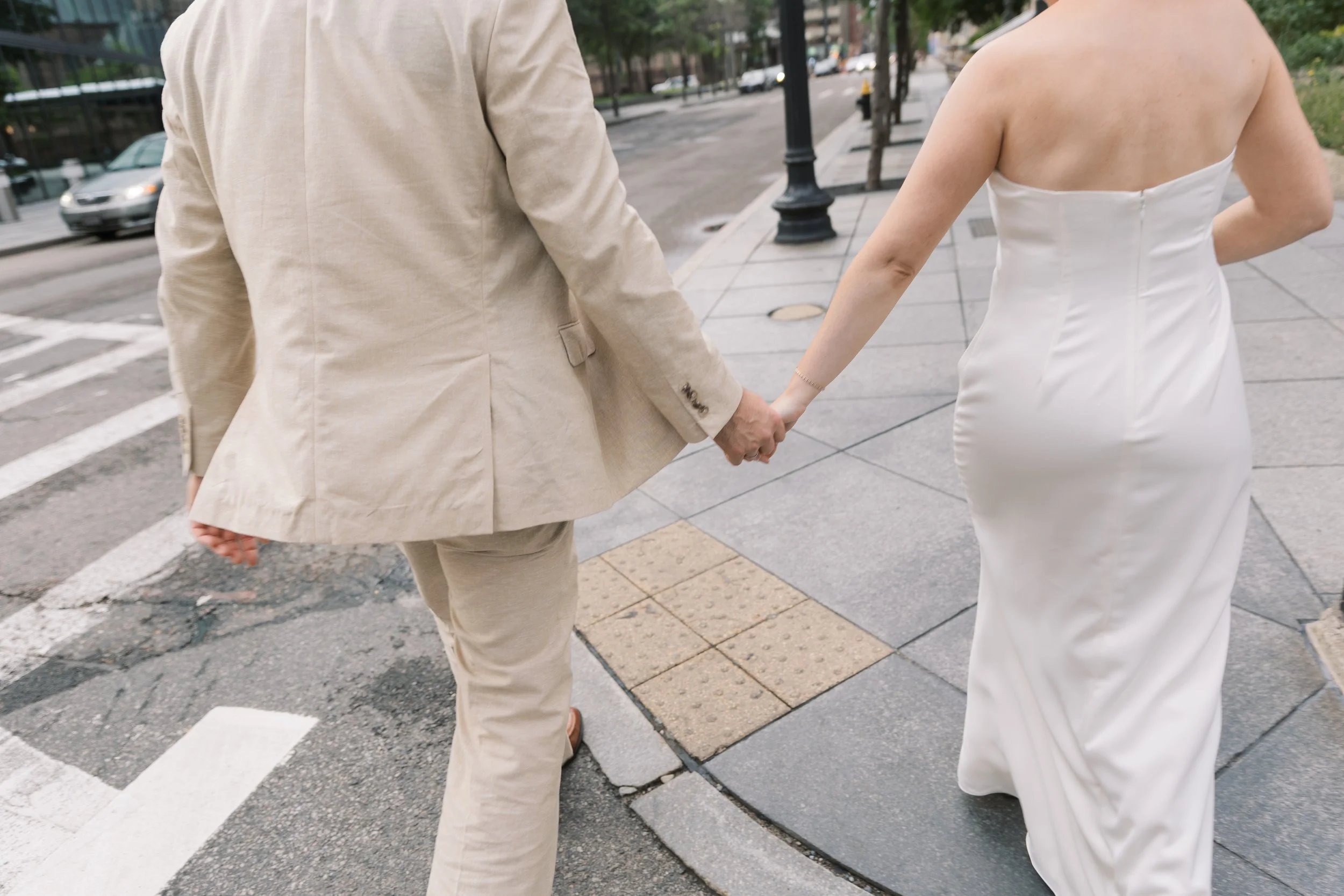 A couple holding hands while walking on a city sidewalk, with one person in a beige suit and the other in a white strapless dress.