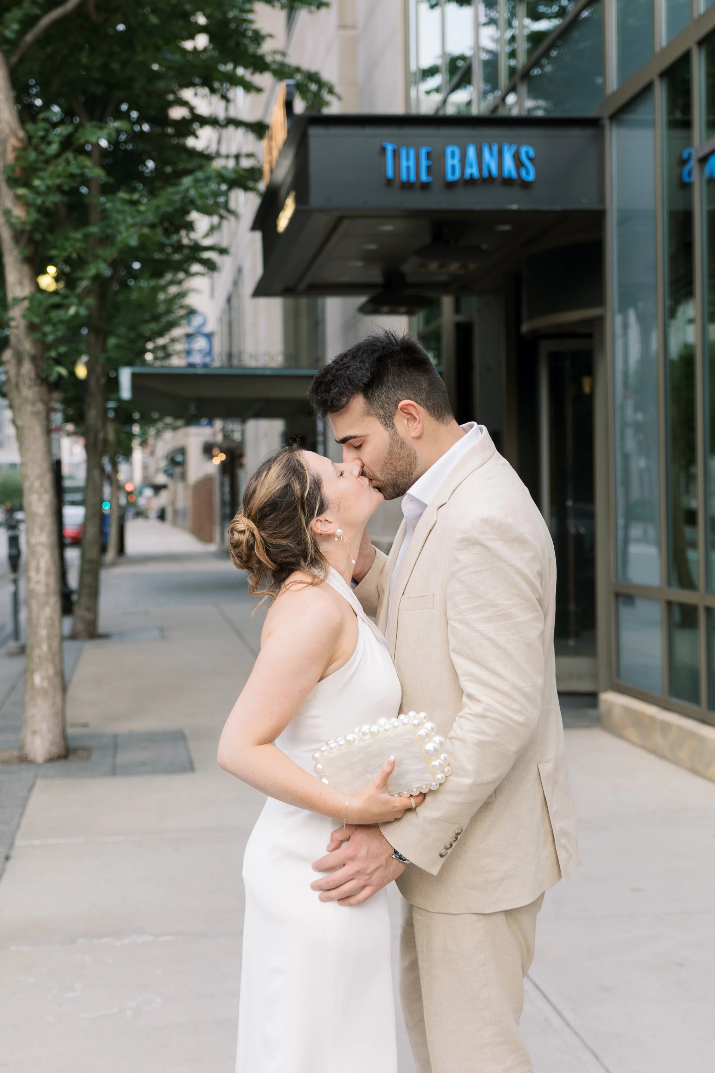 A couple dressed in wedding attire kissing outside a building with a sign that reads 'The Banks'