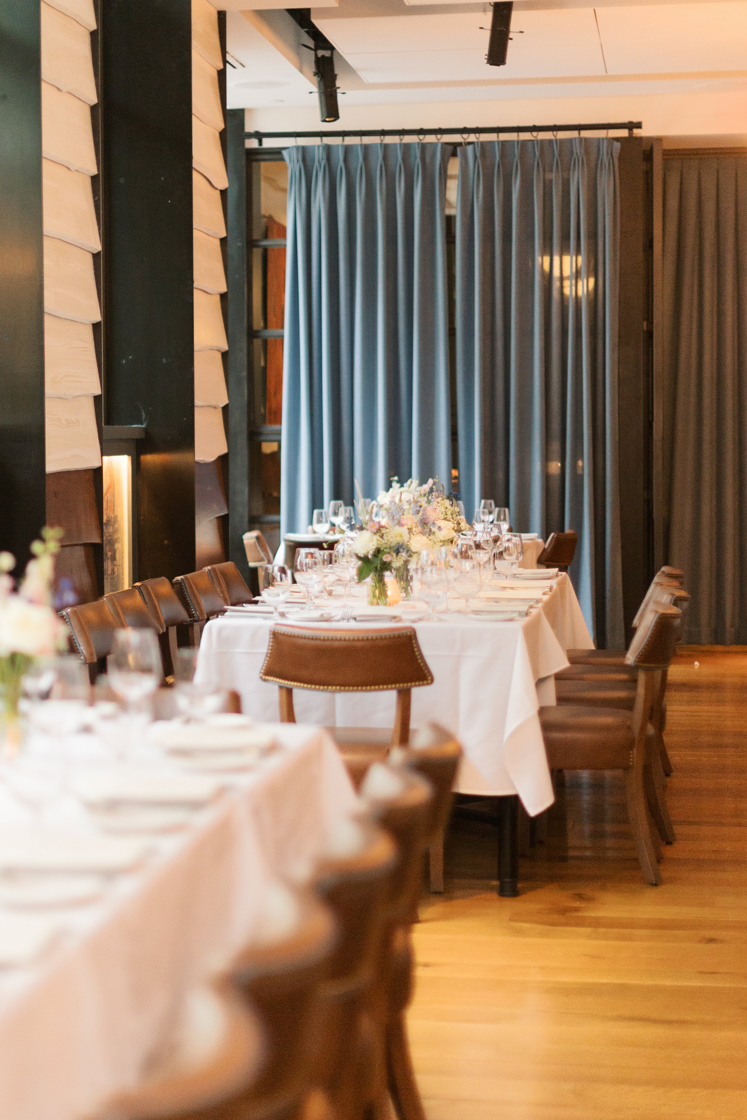 Elegant dining room with round tables covered with white tablecloths, set with wine glasses, plates, and floral centerpieces, with blue curtains and wood-paneled walls.