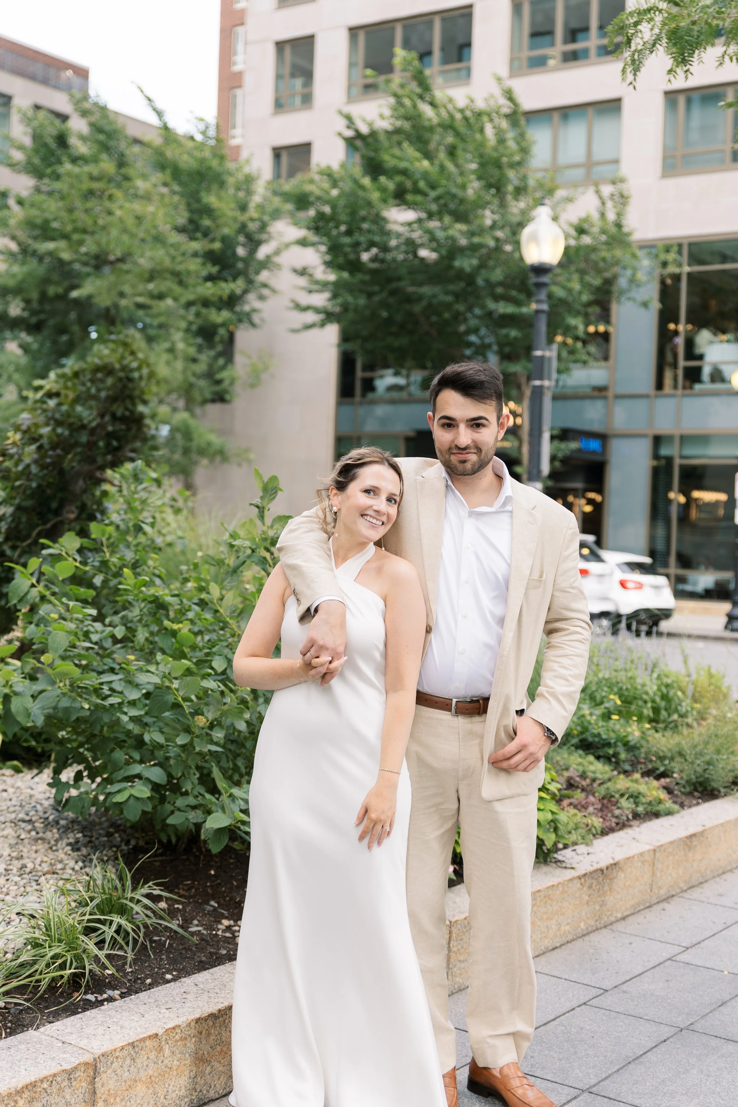 A smiling woman in a white dress and a man in a beige suit with a white shirt, standing outdoors on a city sidewalk with greenery and modern buildings in the background.