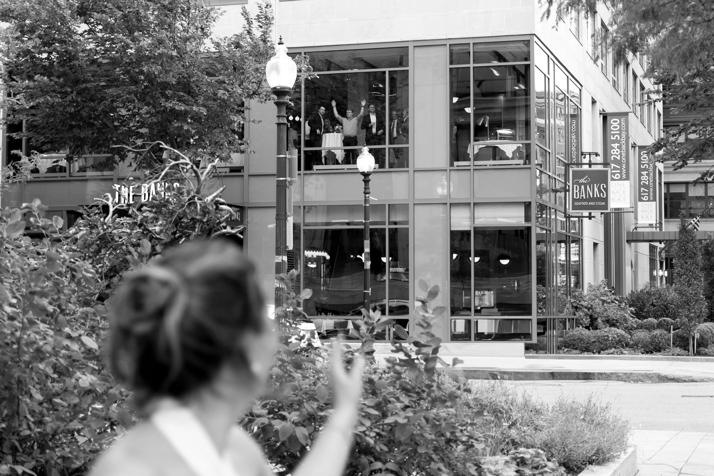 A woman with a blurred face and hair tied up in a bun, pointing towards a building with large glass windows. Inside, on the second floor, a group of four people are seen celebrating, with two of them raising their arms.