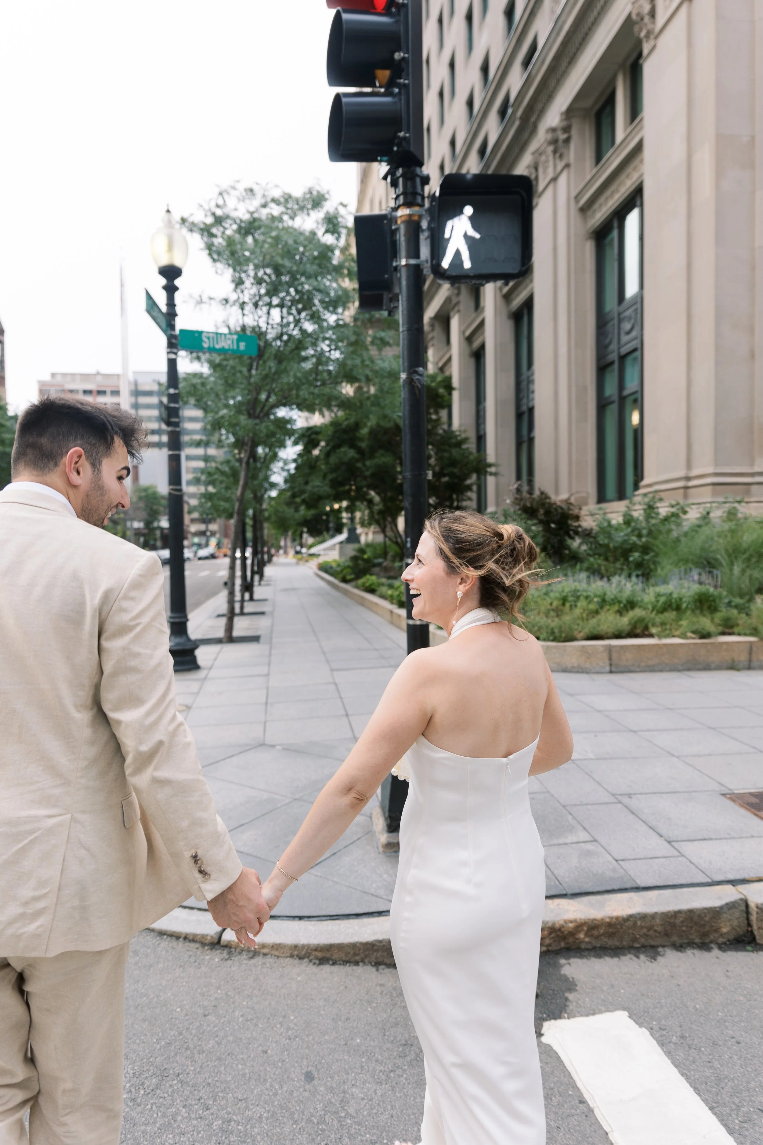 A couple in wedding attire crossing a city street while holding hands, with a pedestrian crossing signal and city buildings in the background.