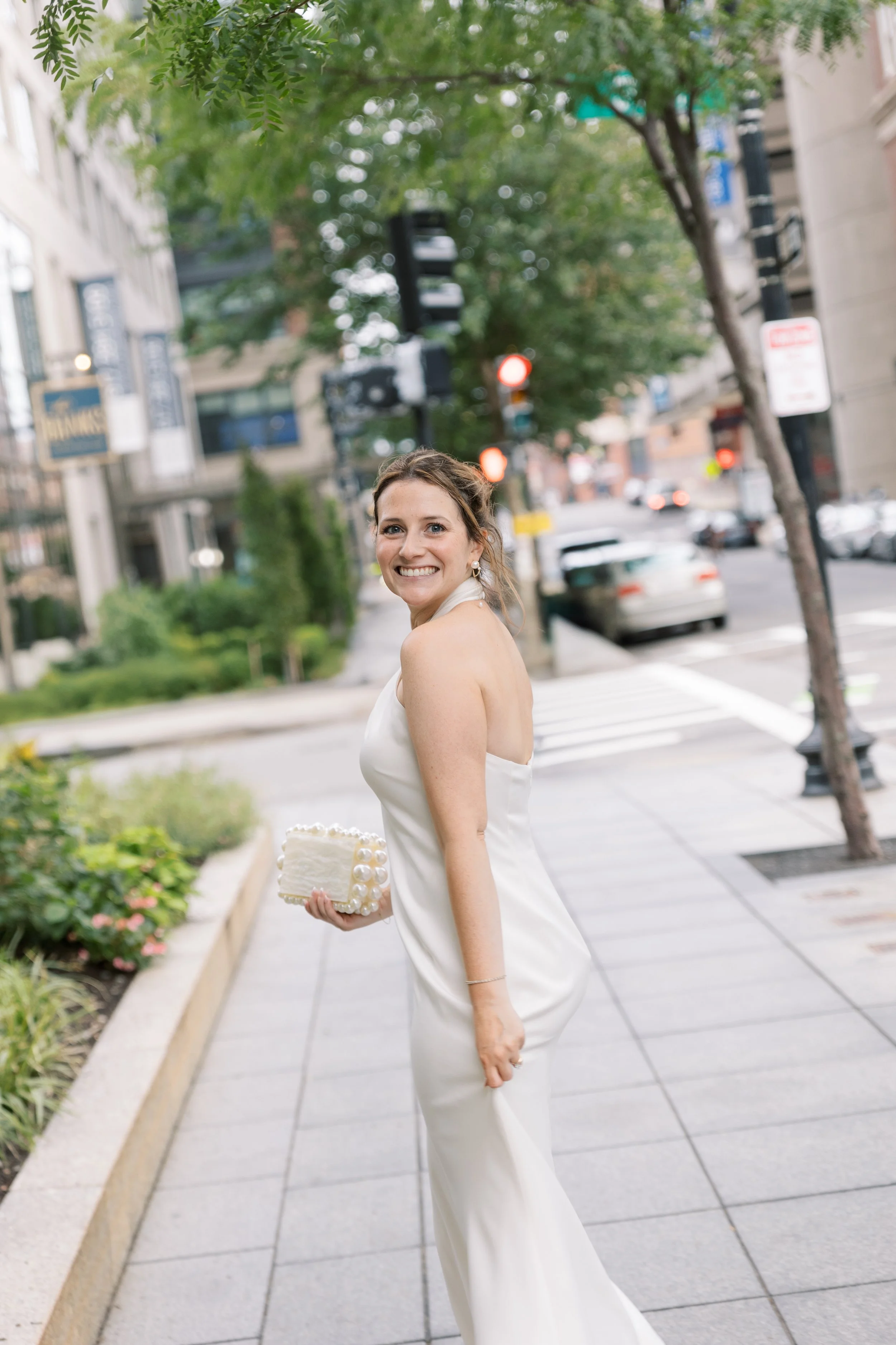 A smiling woman in a white dress holding a small white cake with pearls, standing on a city sidewalk with trees and cars in the background.
