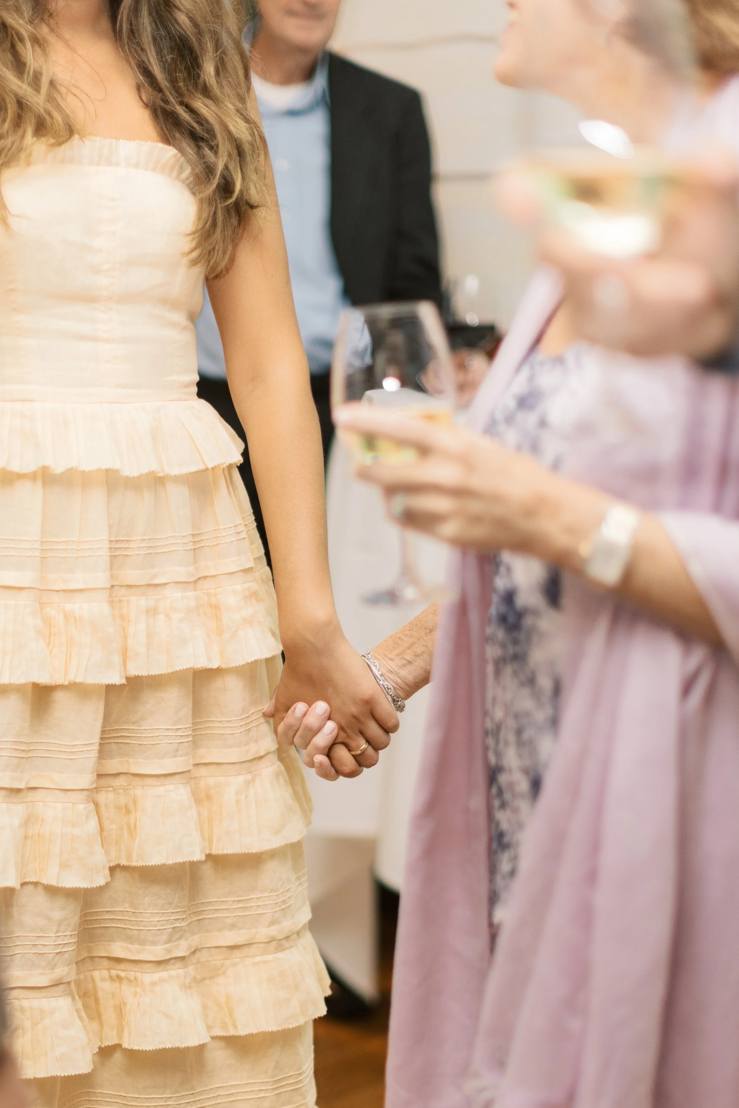 Two women holding hands at a social gathering, with other people in the background, some holding drinks.