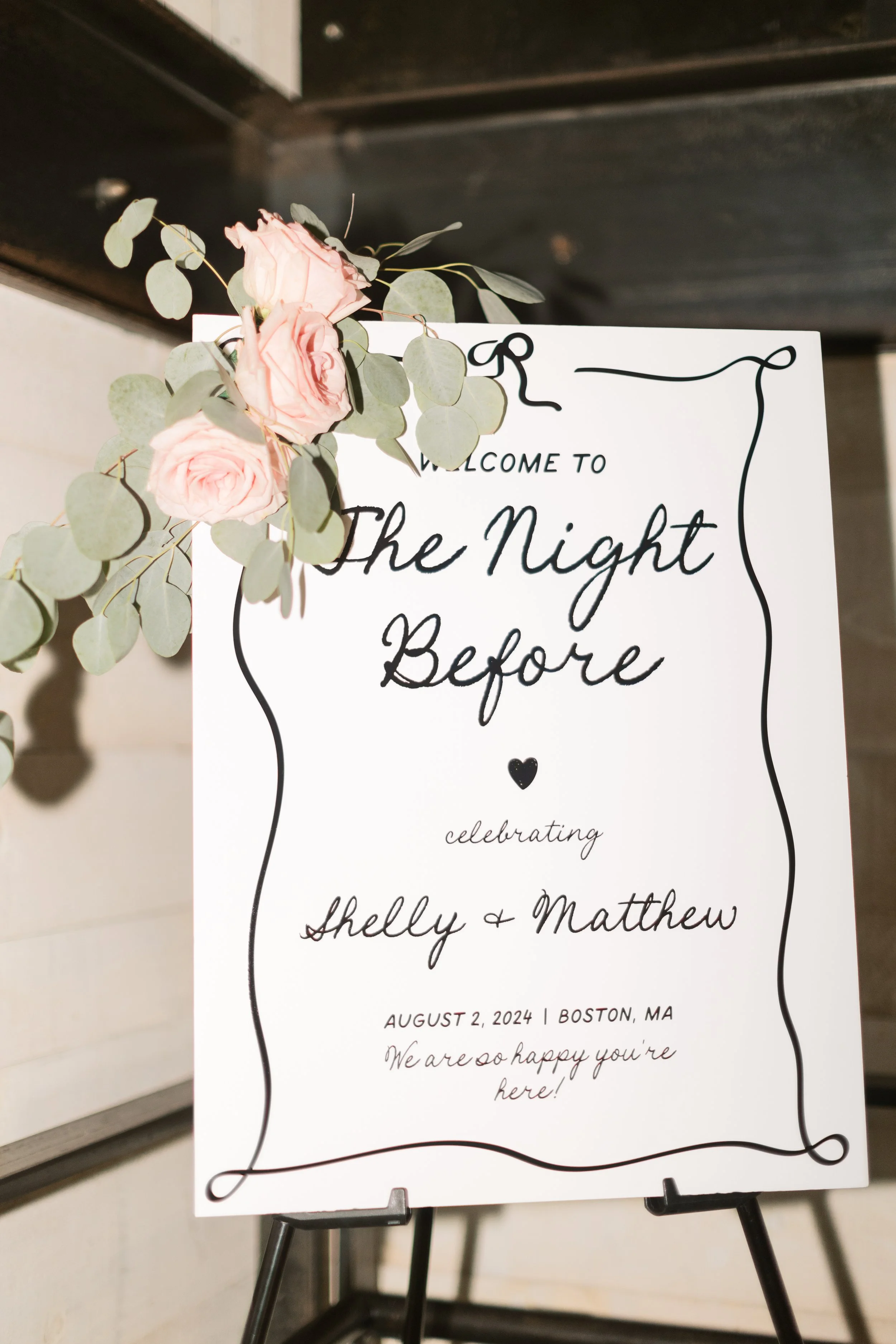 Wedding welcome sign with pink roses and eucalyptus leaves, displaying the event details for Shelly and Matthew's celebration on August 2, 2024, in Boston, MA.