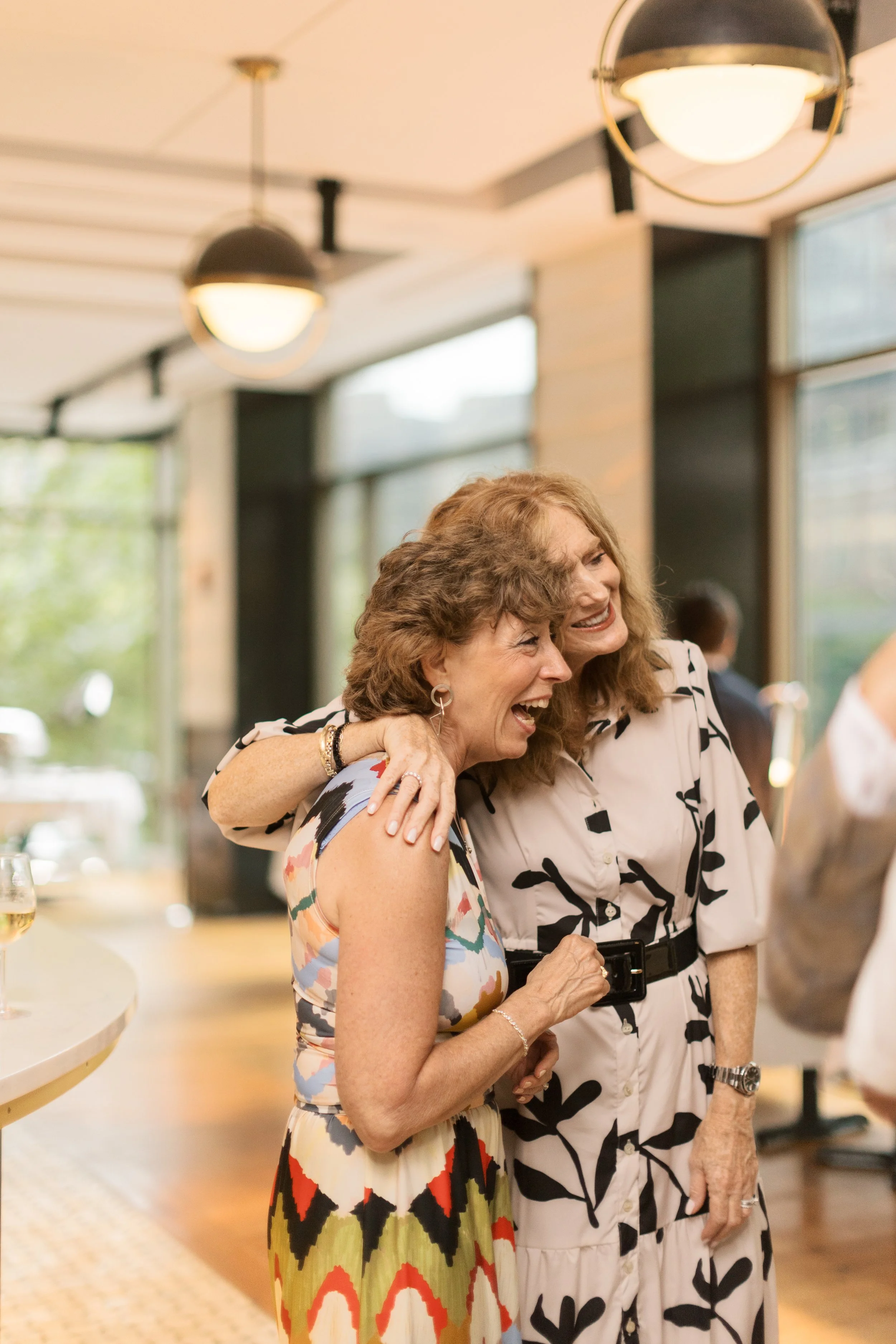 Two women are happily hugging and laughing at a social gathering in a well-lit indoors space with large windows and modern ceiling lights.
