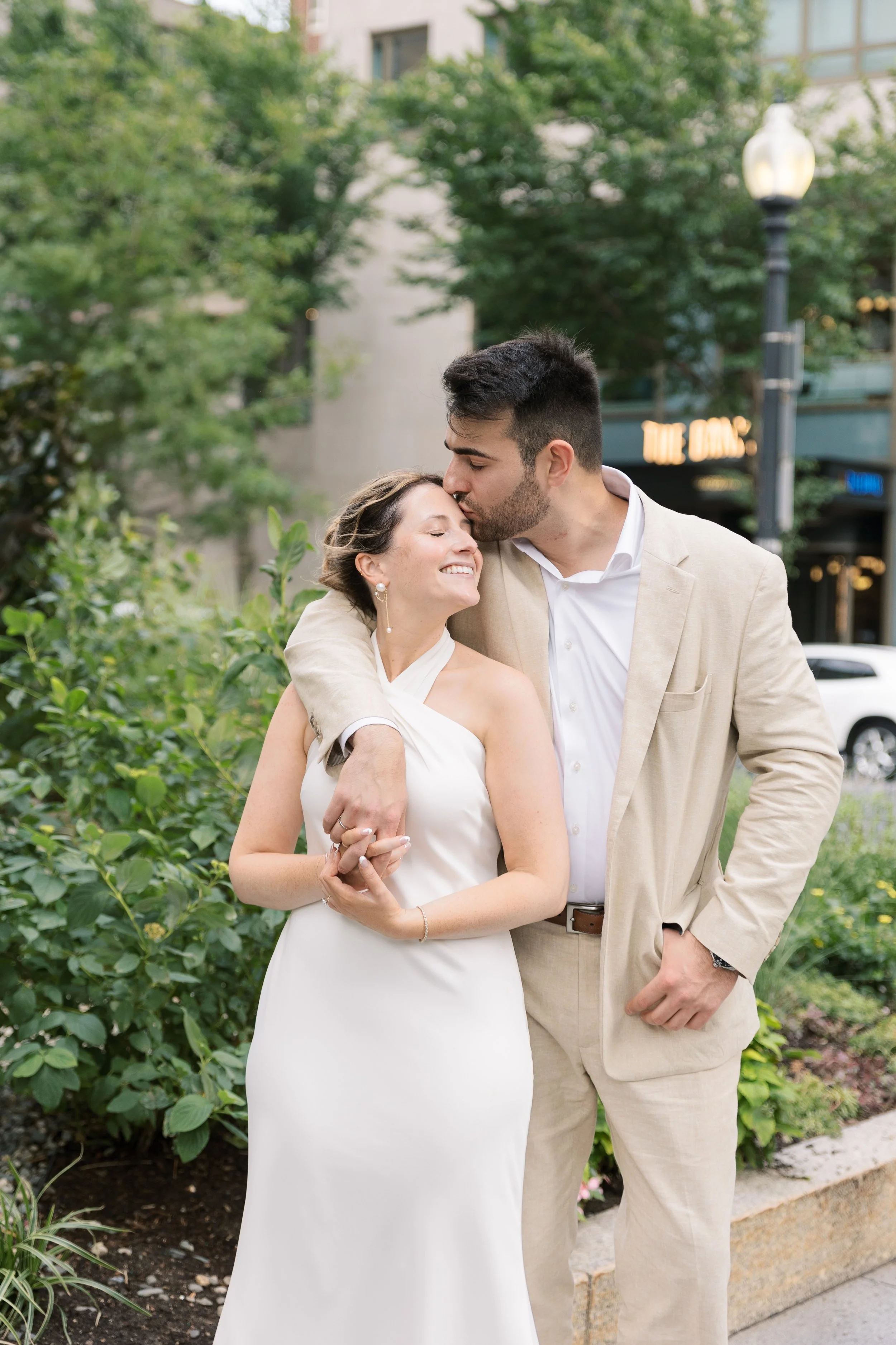 A happy couple in formal attire sharing an affectionate moment outside, with trees and a building in the background.