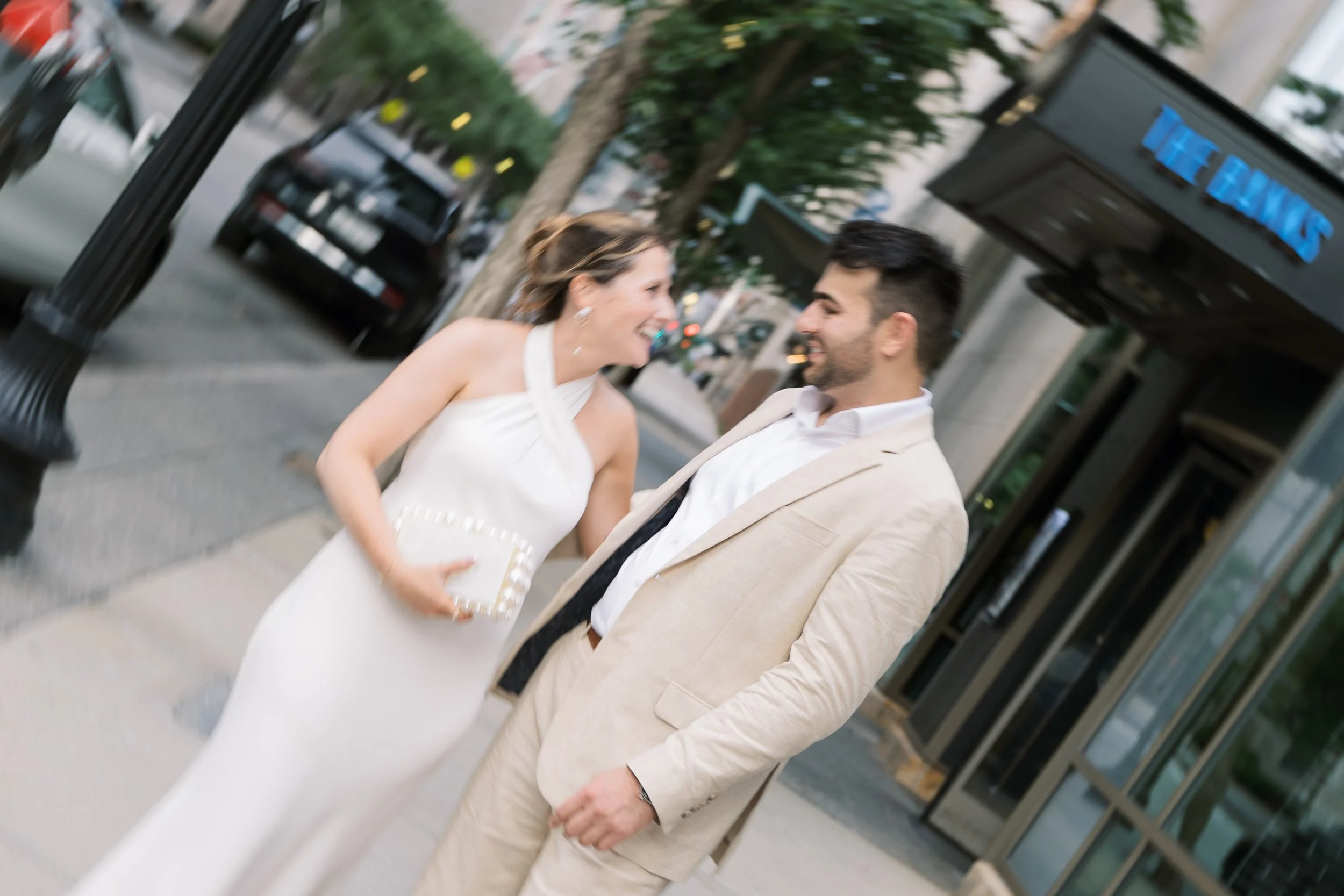 A woman and a man smiling at each other on a city sidewalk, dressed in formal attire, with parked cars and buildings in the background.