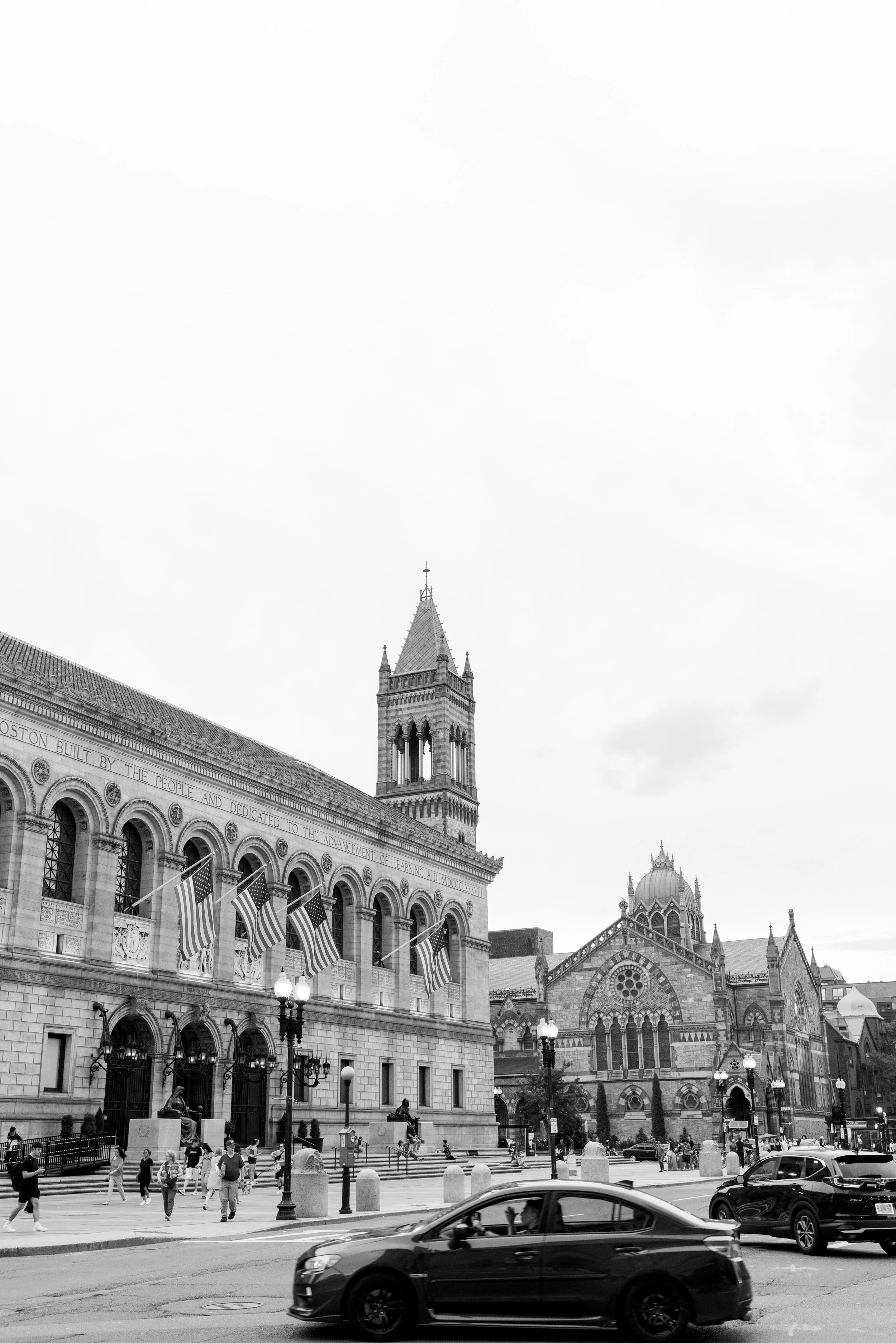 A black and white image of a city street with historic buildings, American flags, pedestrians, and cars.