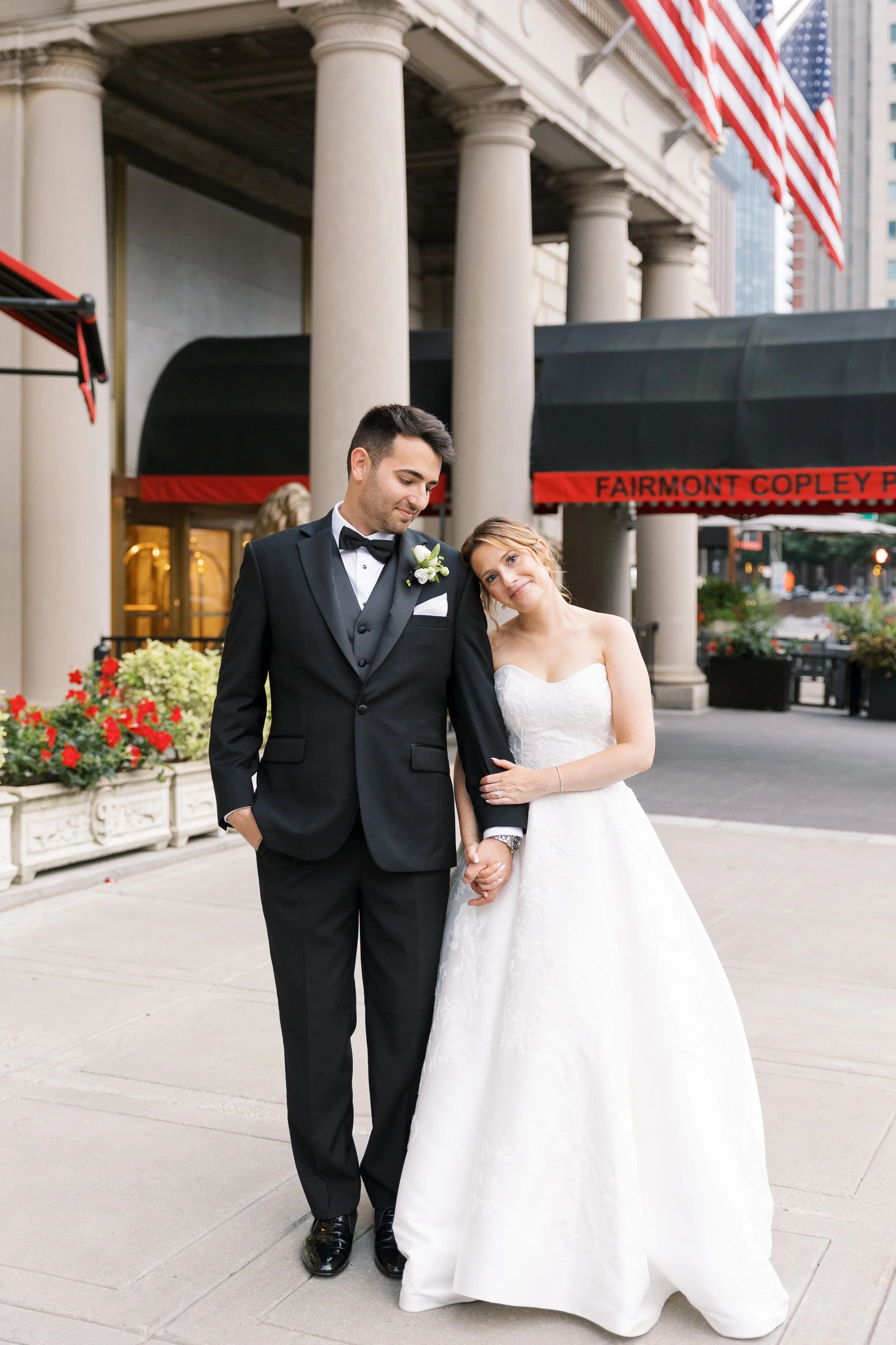 A newlywed couple, dressed in wedding attire, holding hands and standing on a city sidewalk in front of a building with columns and American flags.