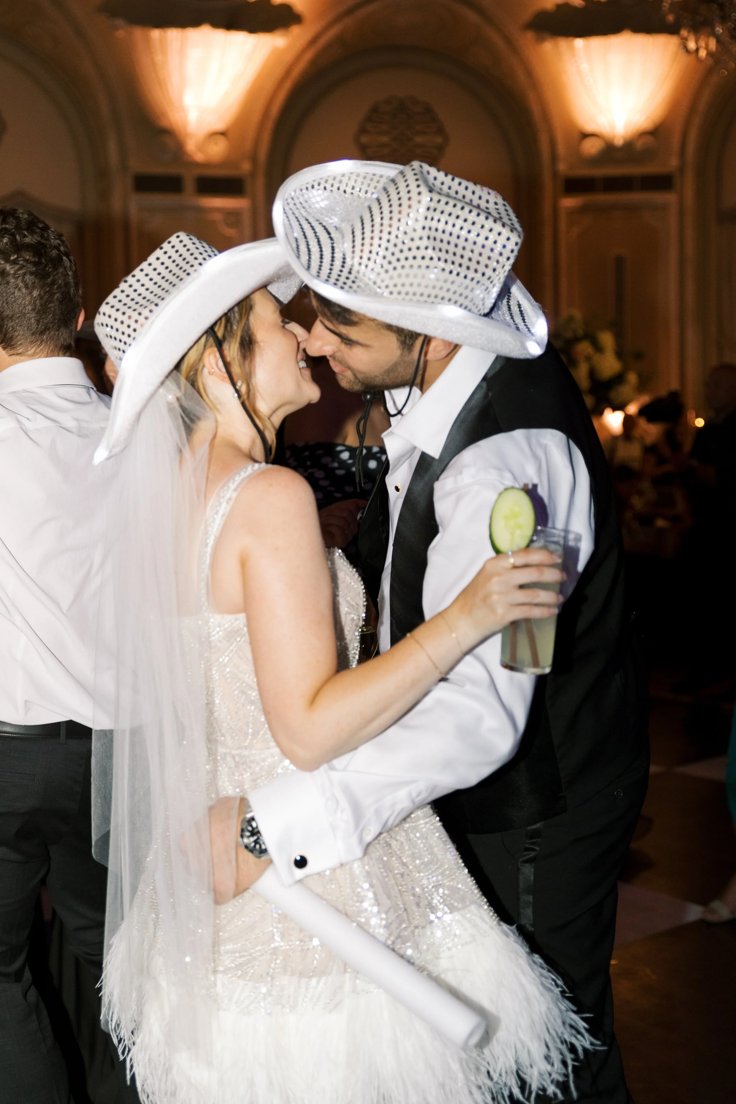 A bride and groom dressed in wedding attire wearing fun, large, white and blue striped cowboy hats, dancing closely at their wedding reception, with a glass of cucumber-infused drink in the bride's hand and the warmly lit event hall in the background