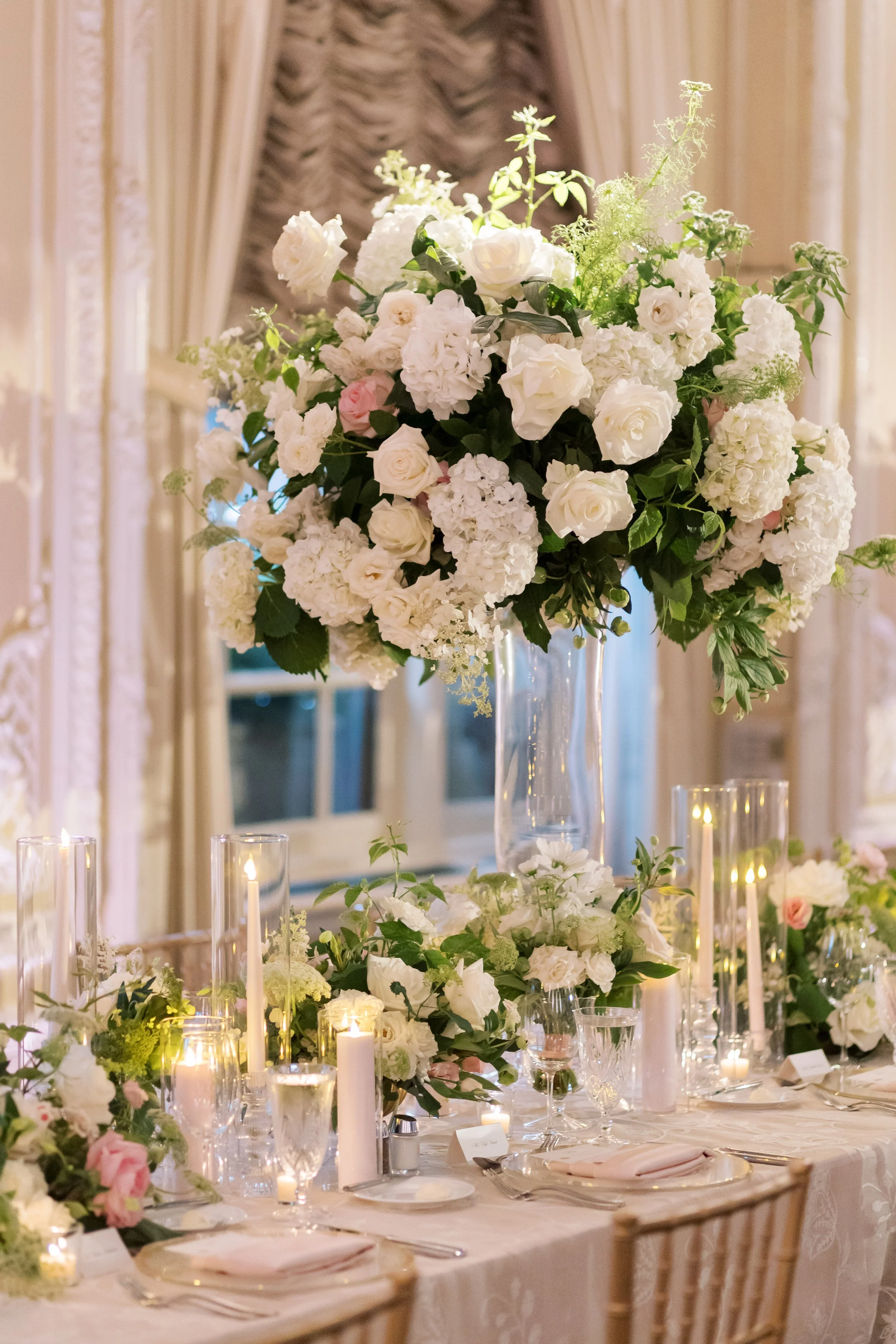 Elegant wedding table centerpiece with white flowers in a tall glass vase and surrounding candles, set in a decorated room.