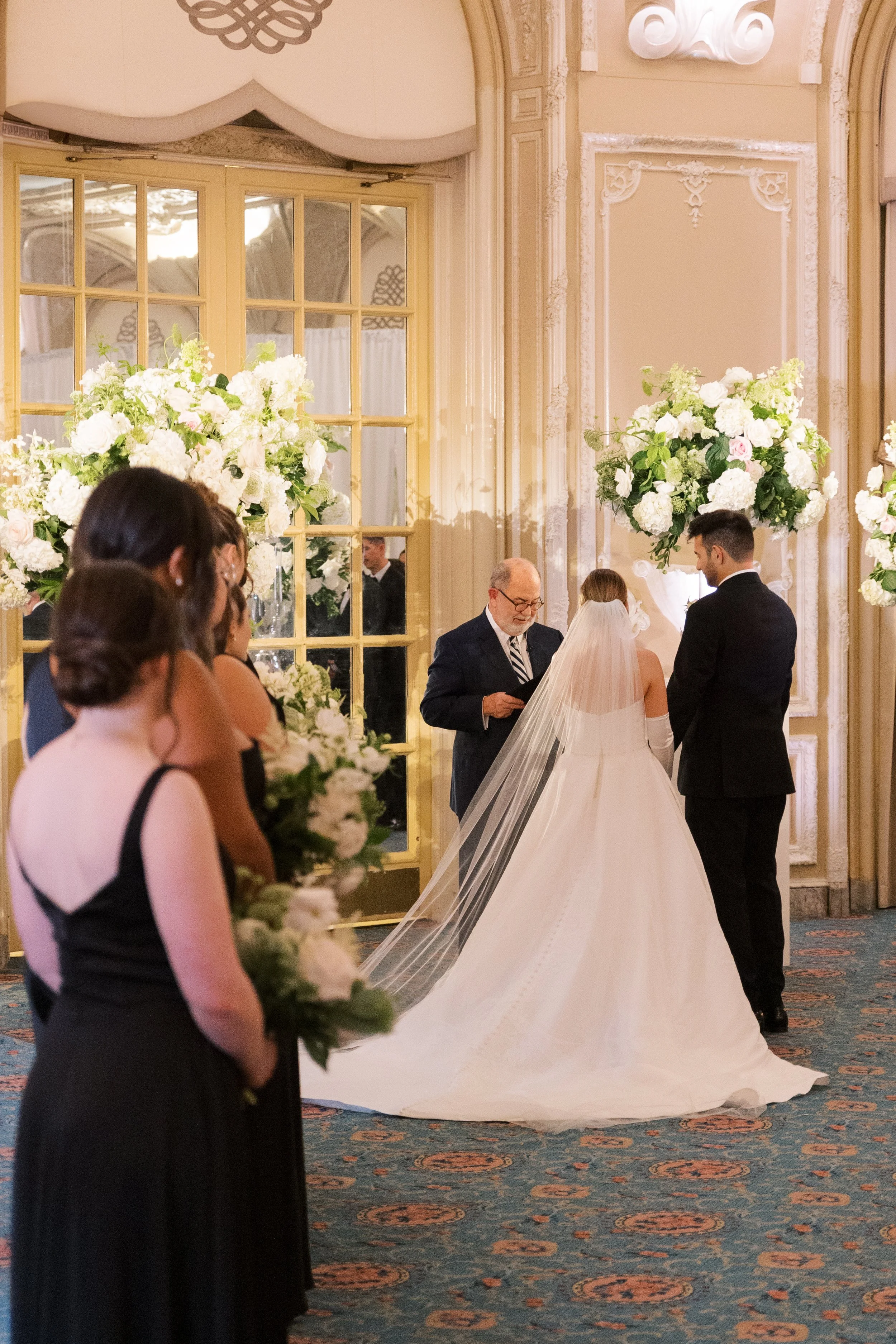 A wedding ceremony with a bride and groom facing an officiant, surrounded by floral arrangements in a decorated hall.