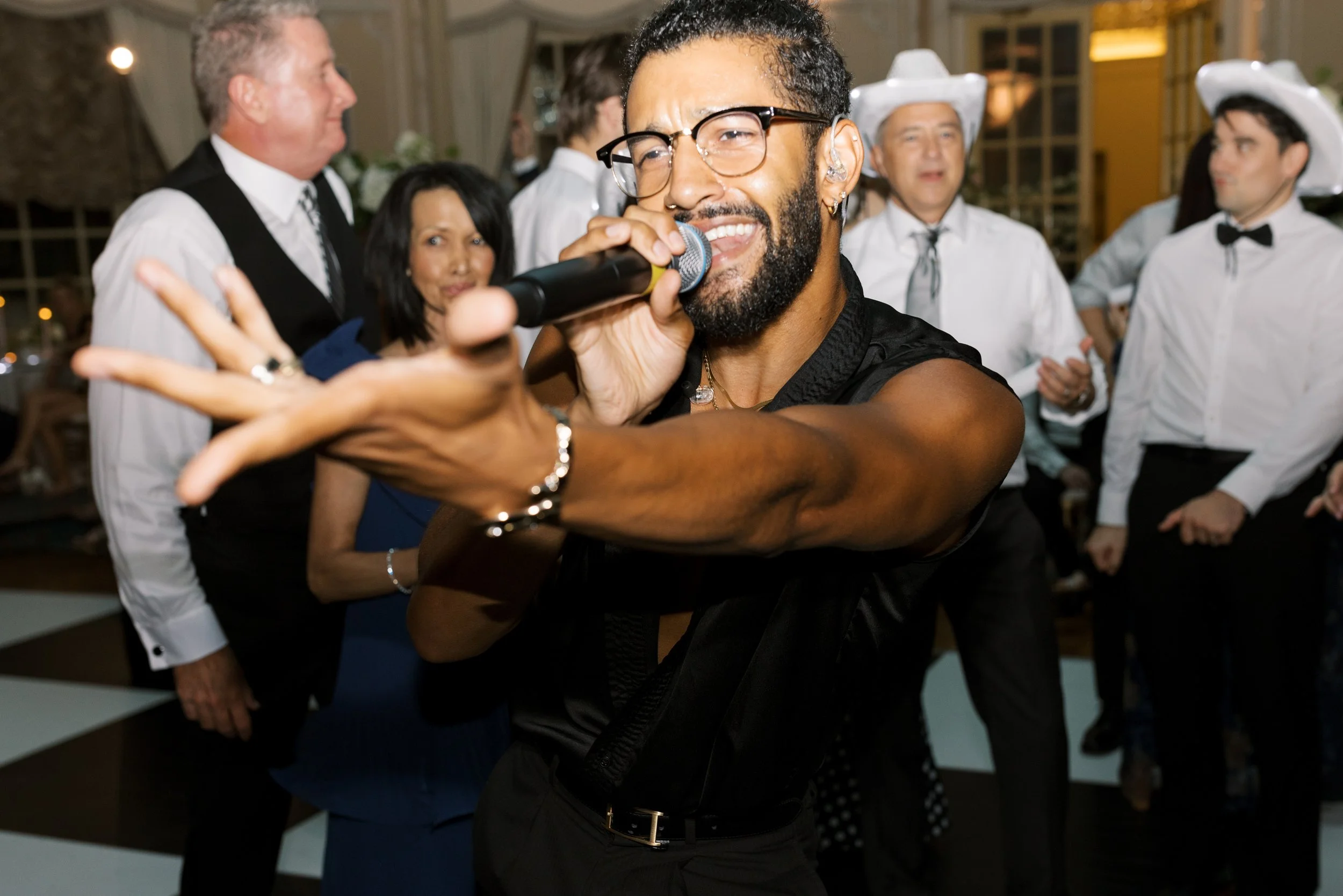 A man with glasses and a beard sings into a microphone at a lively party, with guests in formal and cowboy hats dancing in the background.