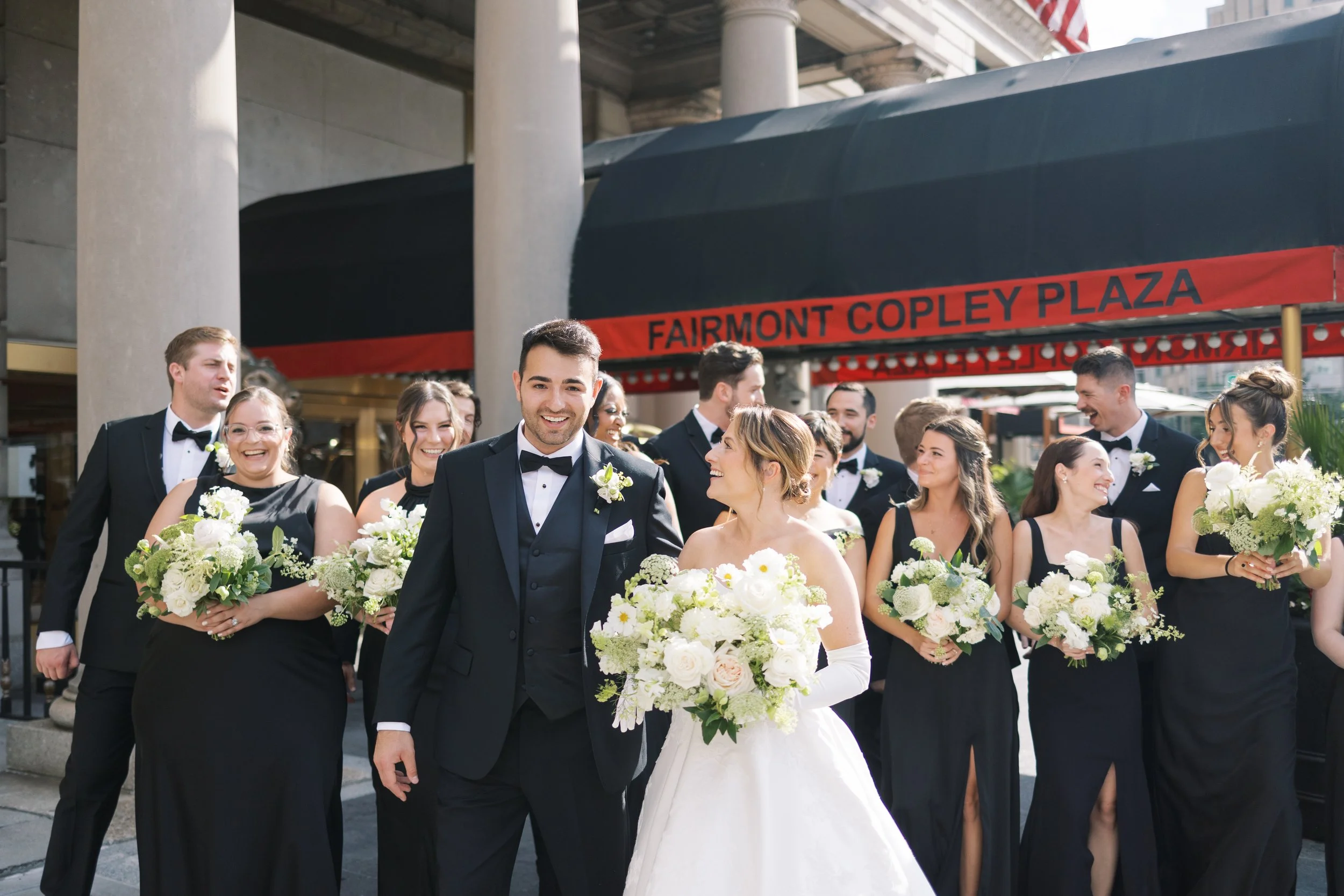 A wedding party outside of Fairmont Copley Plaza hotel, with a bride, groom, and bridesmaids and groomsmen dressed in formal attire, holding bouquets of white and green flowers, under a large black canopy.