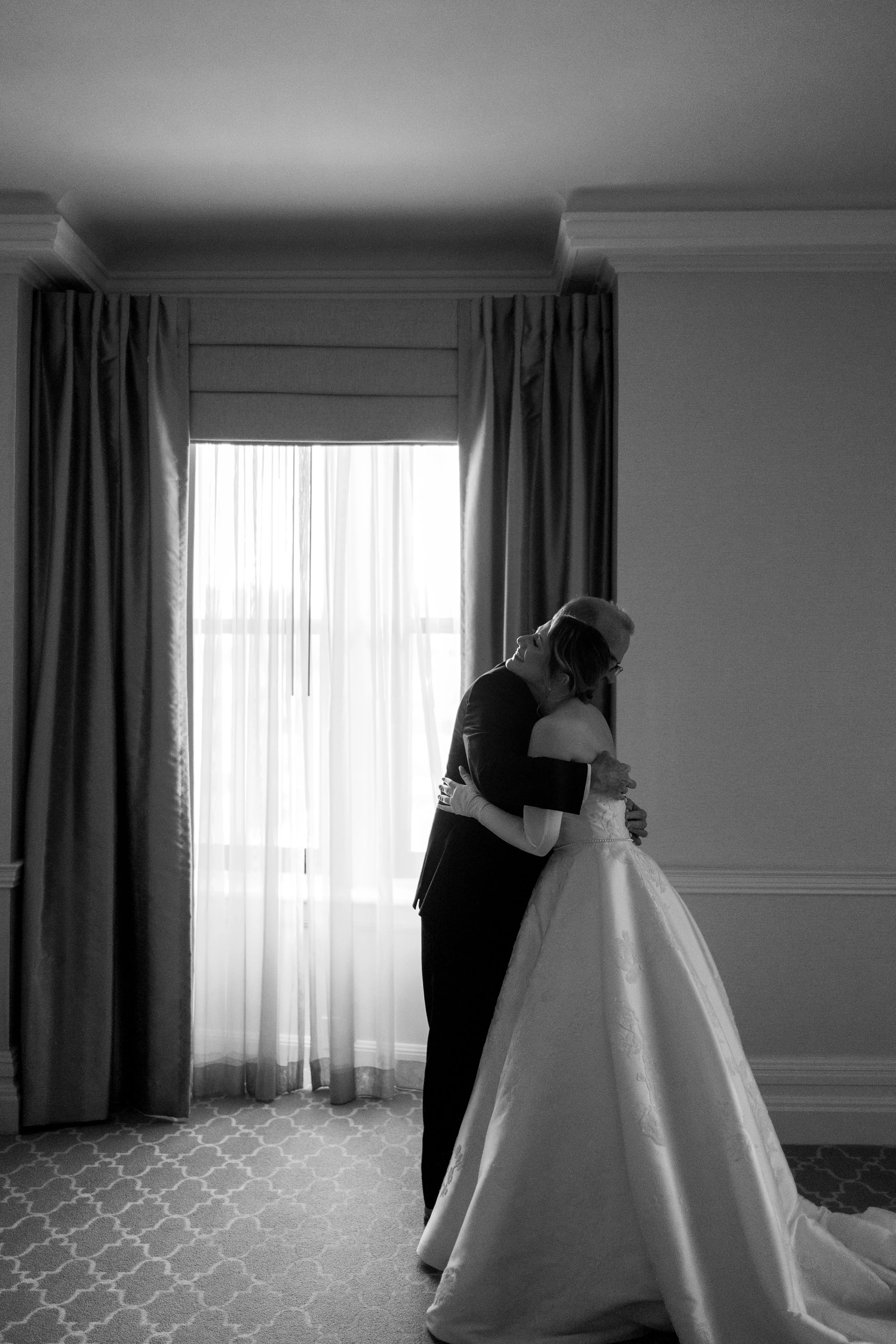 A bride and older man hugging in a hotel room, backlit by a large window with curtains.