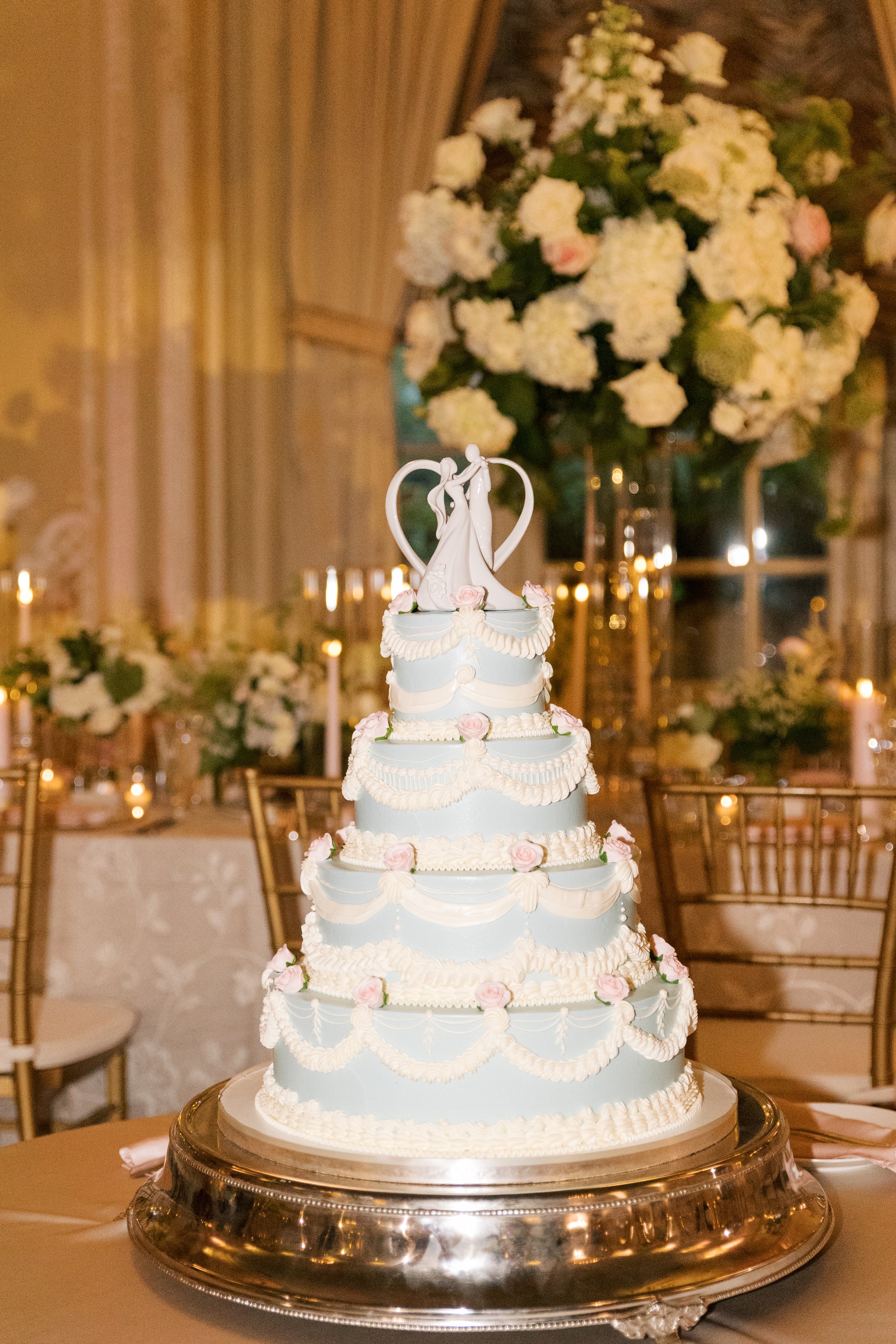A multi-tiered wedding cake decorated with white and pink icing, topped with a figurine of a couple dancing, with a large floral arrangement of white and pink flowers in the background.