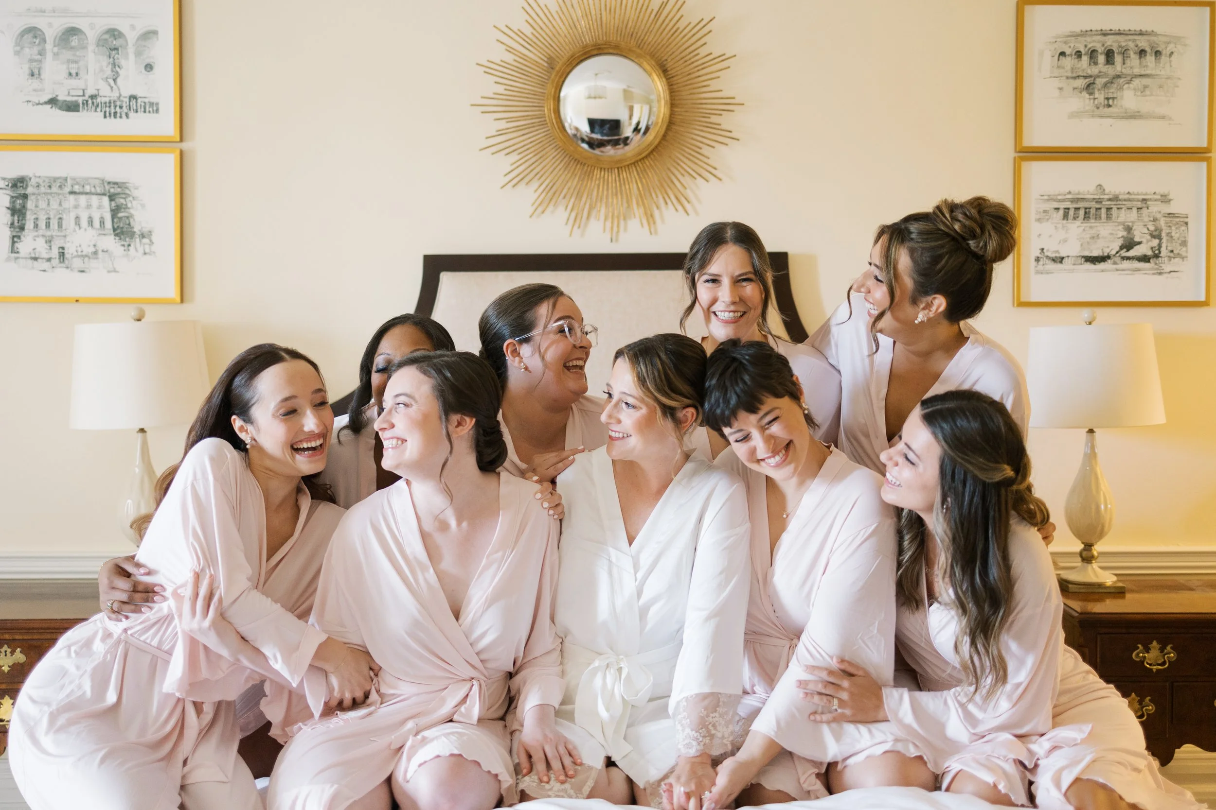 A group of nine women dressed in matching light pink robes, sitting on a bed and smiling happily, sharing a joyful moment together in a cozy room with framed artwork and lamps in the background.