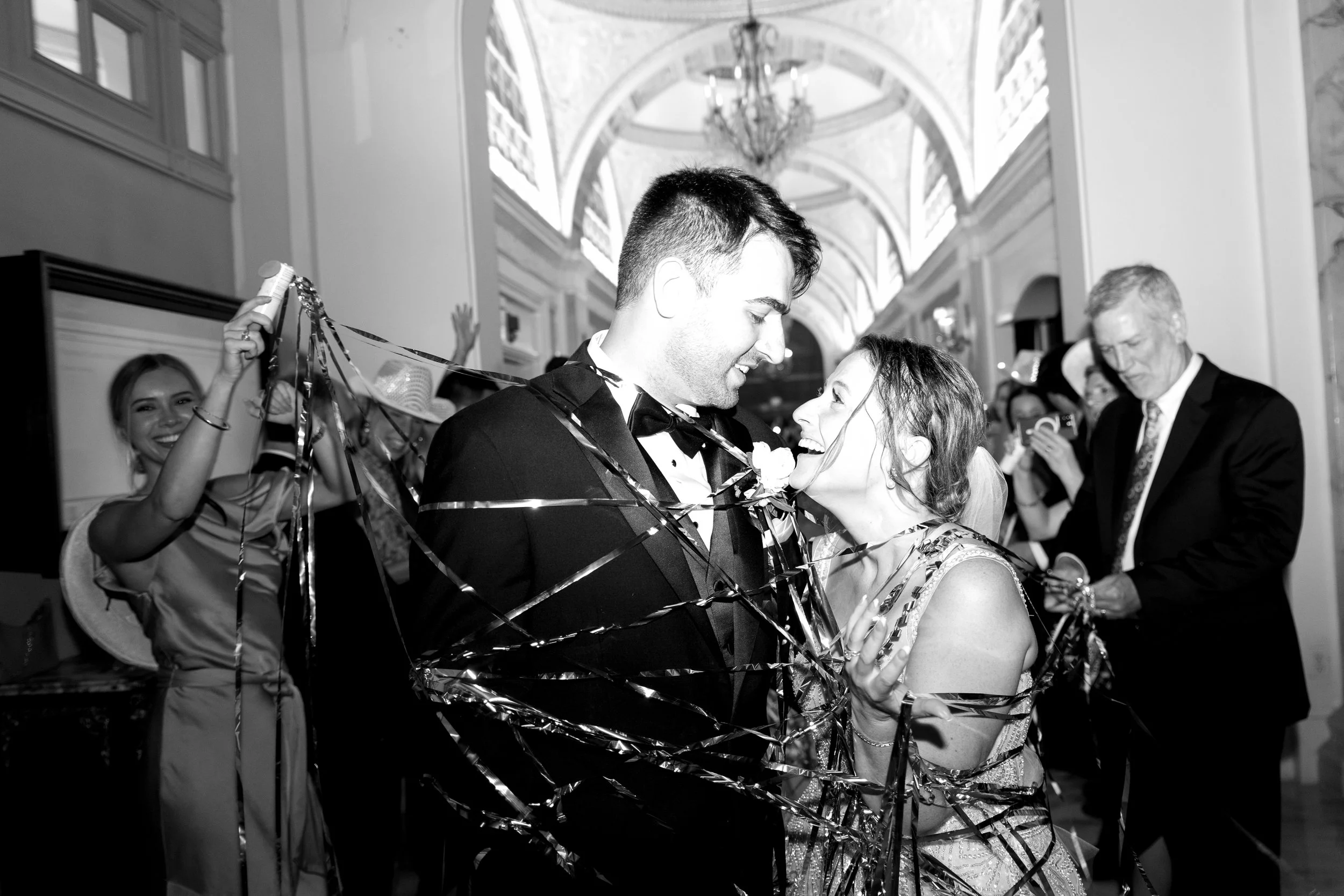 A couple celebrating at a wedding reception, wrapped in streamers, smiling at each other, with guests around them taking photos and cheering.