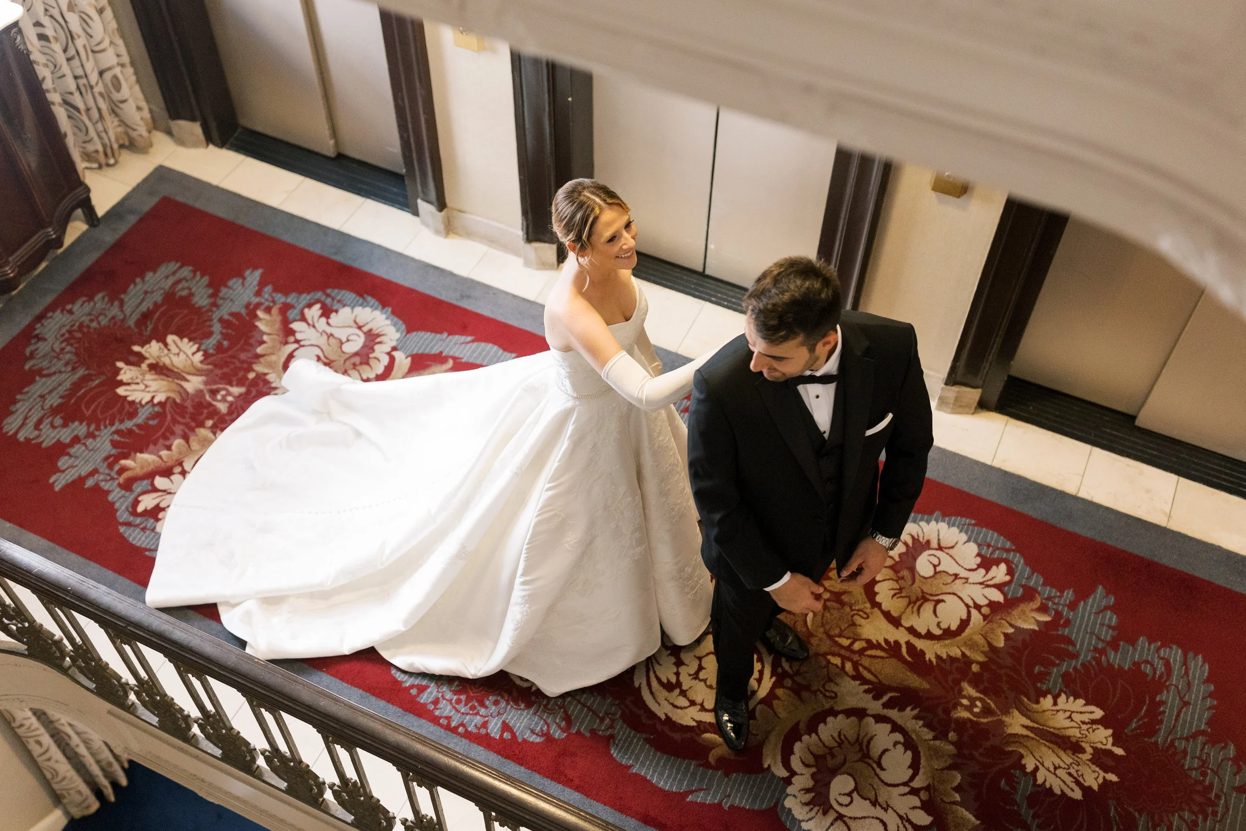 Bride and groom on a staircase, with the bride smiling and touching the groom's shoulder, both dressed in wedding attire.