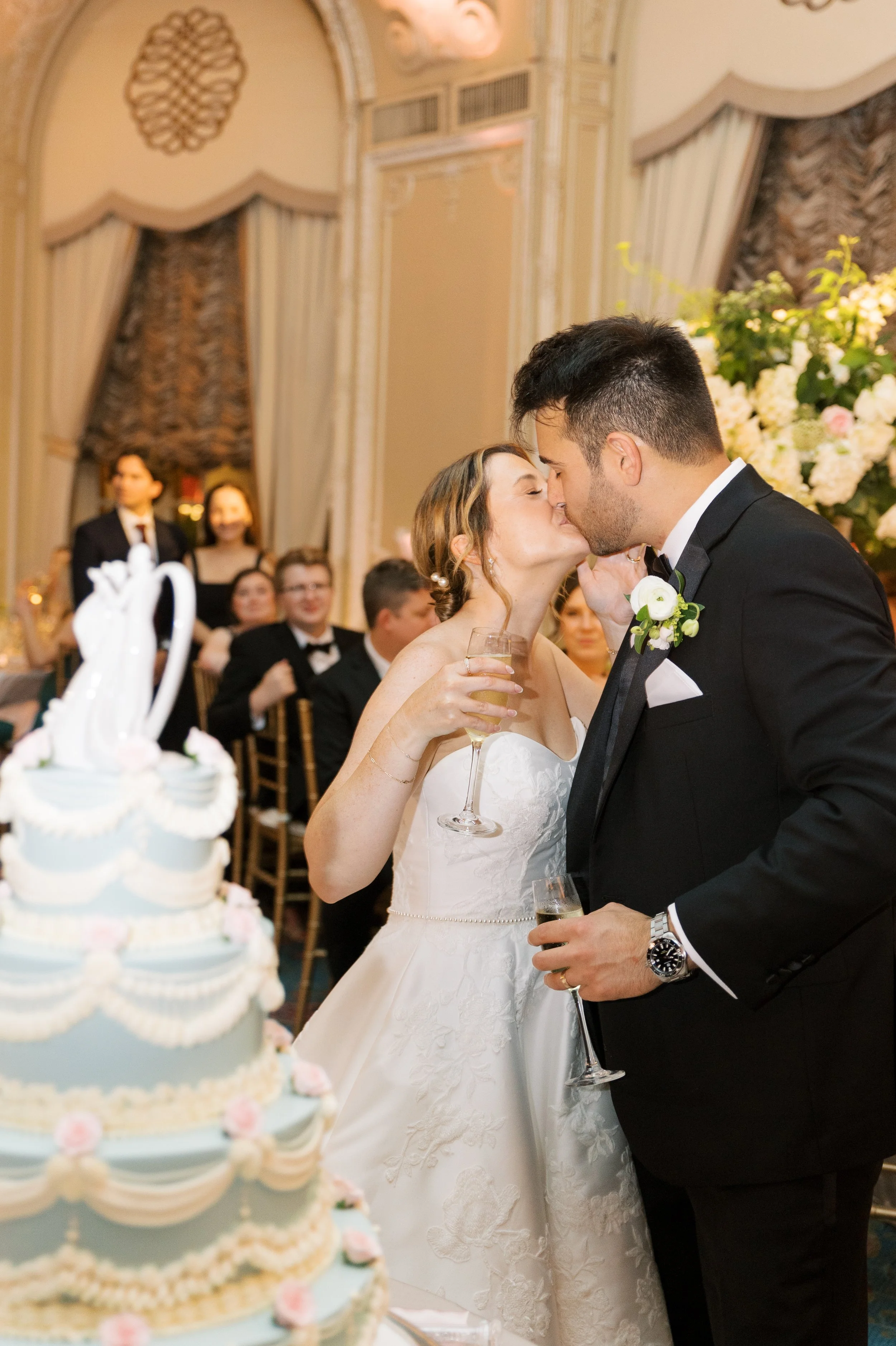A bride and groom share a kiss at their wedding reception, holding champagne glasses. The bride is in a white gown and the groom in a black tuxedo. A wedding cake decorated with pearls and pink roses is in the foreground, and guests are seated in the