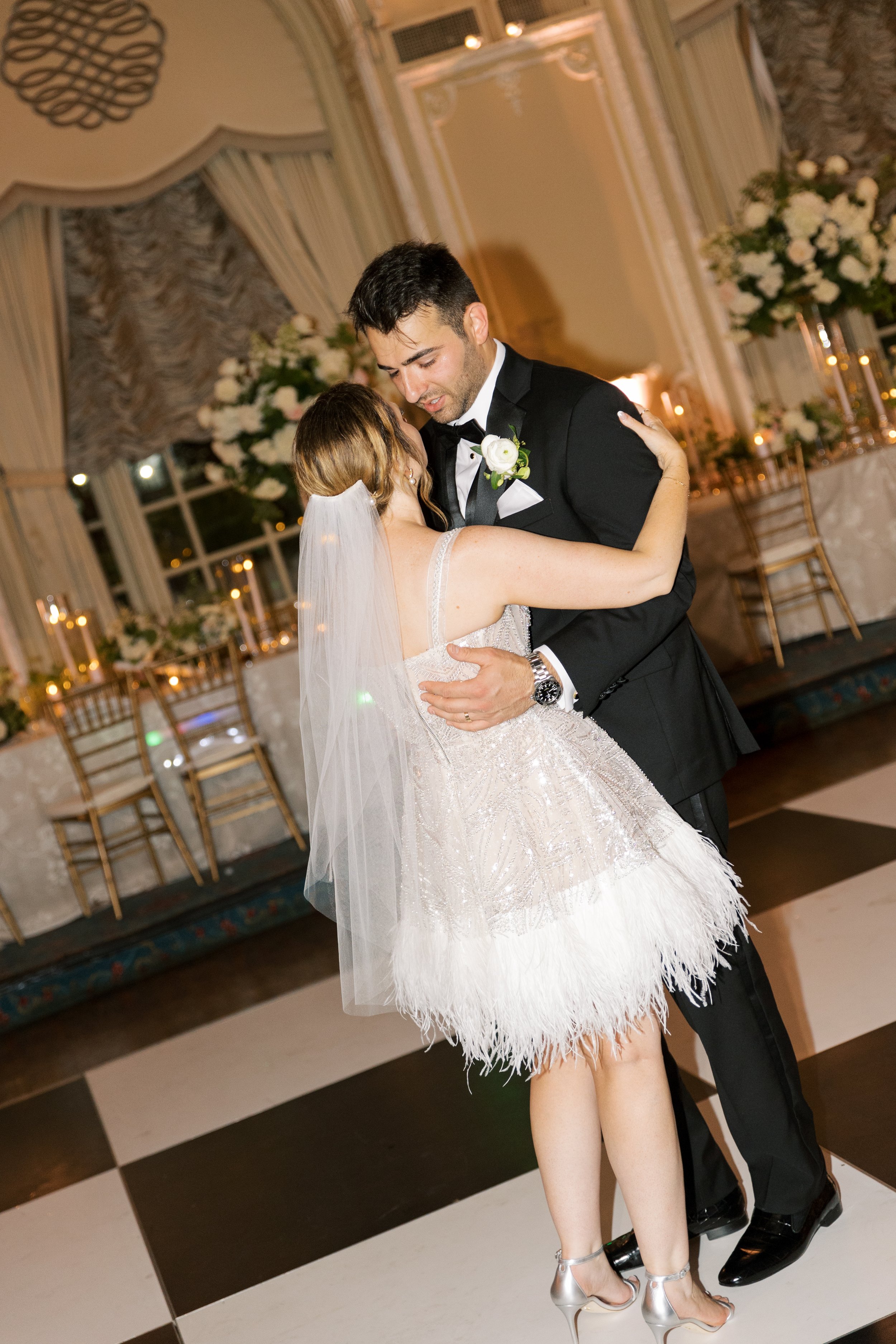 A bride and groom are dancing closely at their wedding reception, sharing an intimate moment on the dance floor. The bride is wearing a short, bridal dress with feather embellishments and a veil, while the groom is dressed in a black tuxedo with a wh
