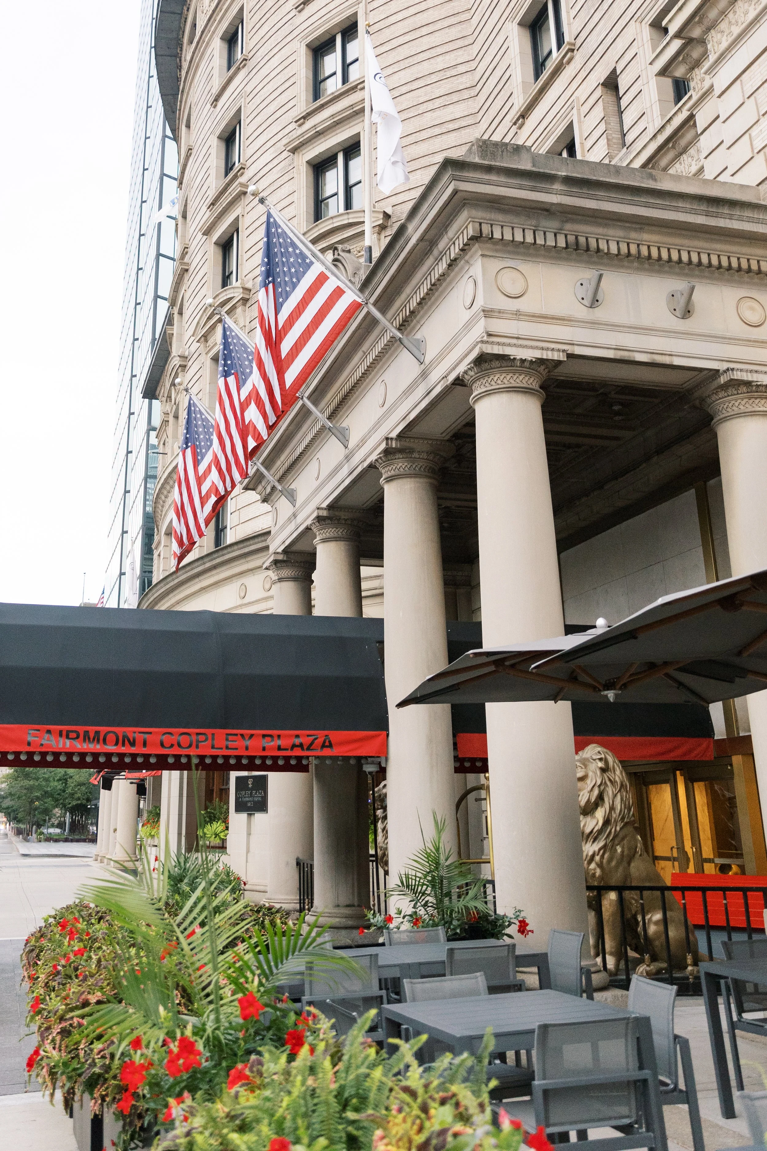 Facade of a building with American flags, outdoor seating area with tables and chairs, and potted plants at Fairmont Copley Plaza hotel.