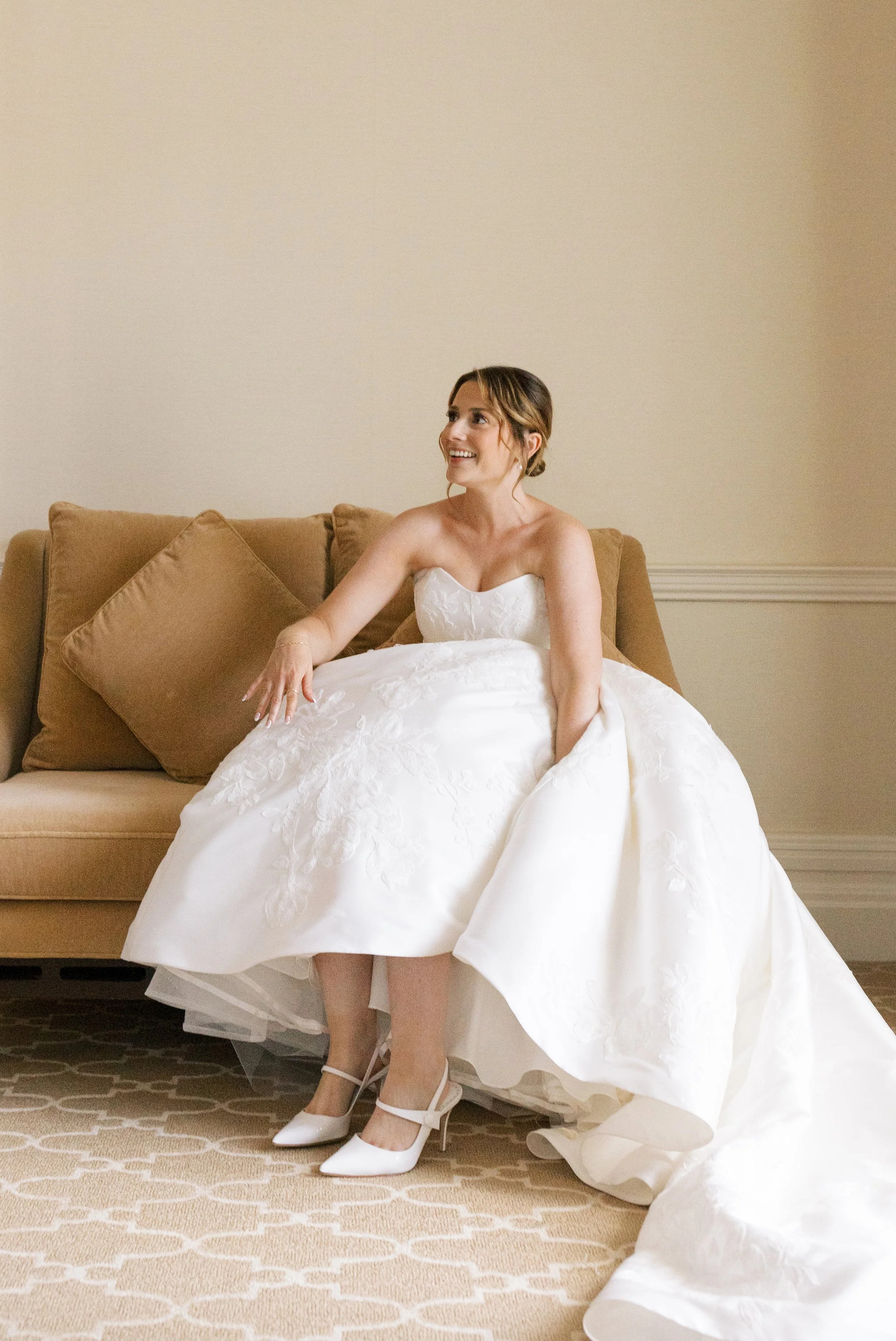 A bride in a strapless white wedding gown sitting on a beige sofa with brown pillows, smiling and looking to her left, wearing white heels.